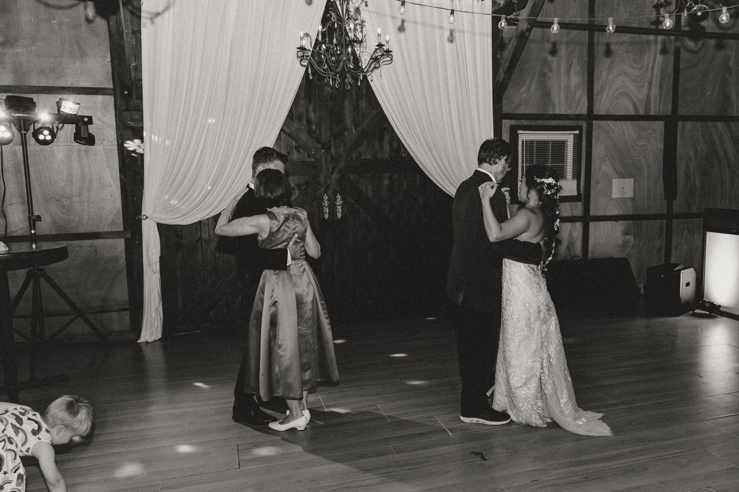 Black and white photo of couples sharing first dance at elegant wedding reception with draped curtains and chandelier.