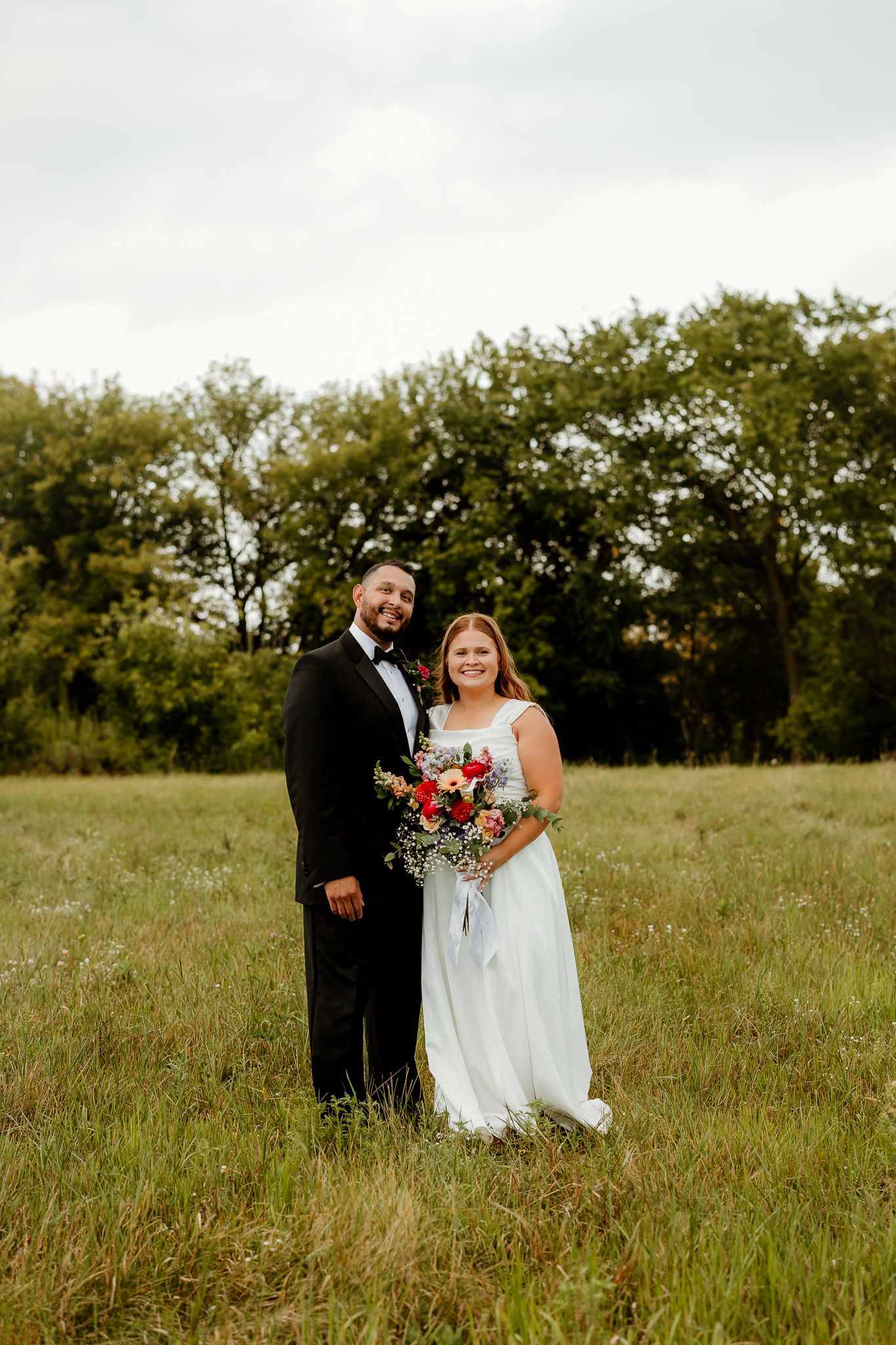 A bride and groom pose in a lush green field, she holds a red and white bouquet, he wears a black tuxedo.