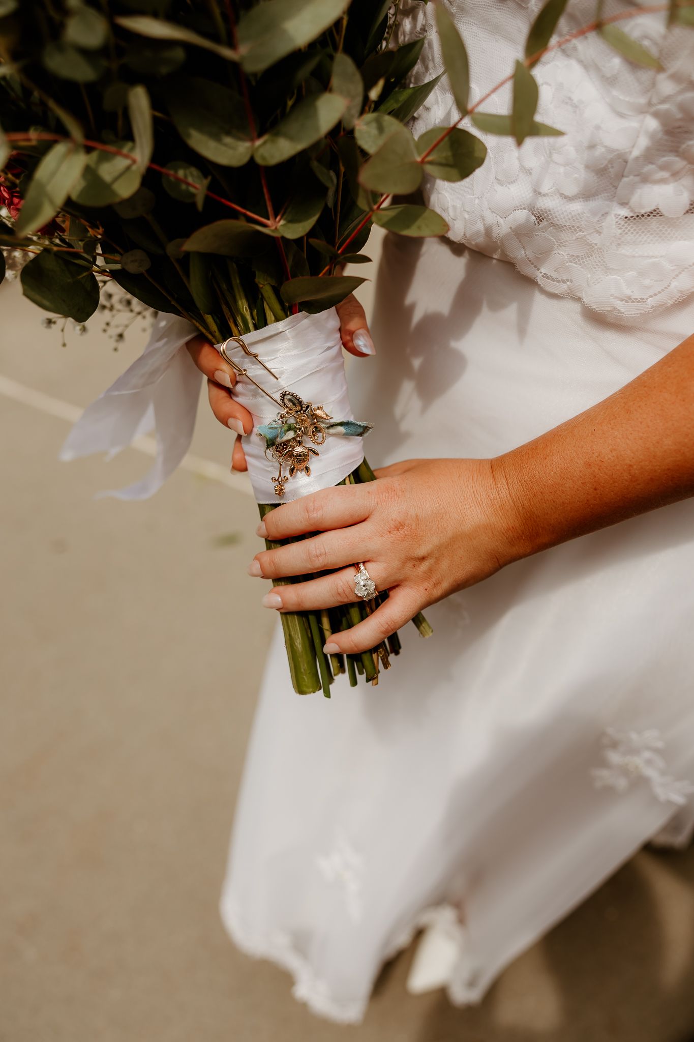 Bride holding a wedding bouquet with eucalyptus and a diamond engagement ring, wrapped with white ribbon and floral charm.