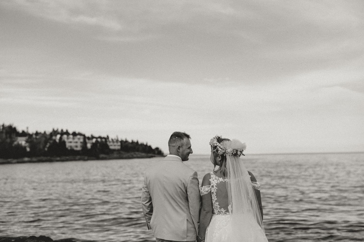 Black and white photo of a bride and groom standing by the ocean, gazing at the water on their wedding day.