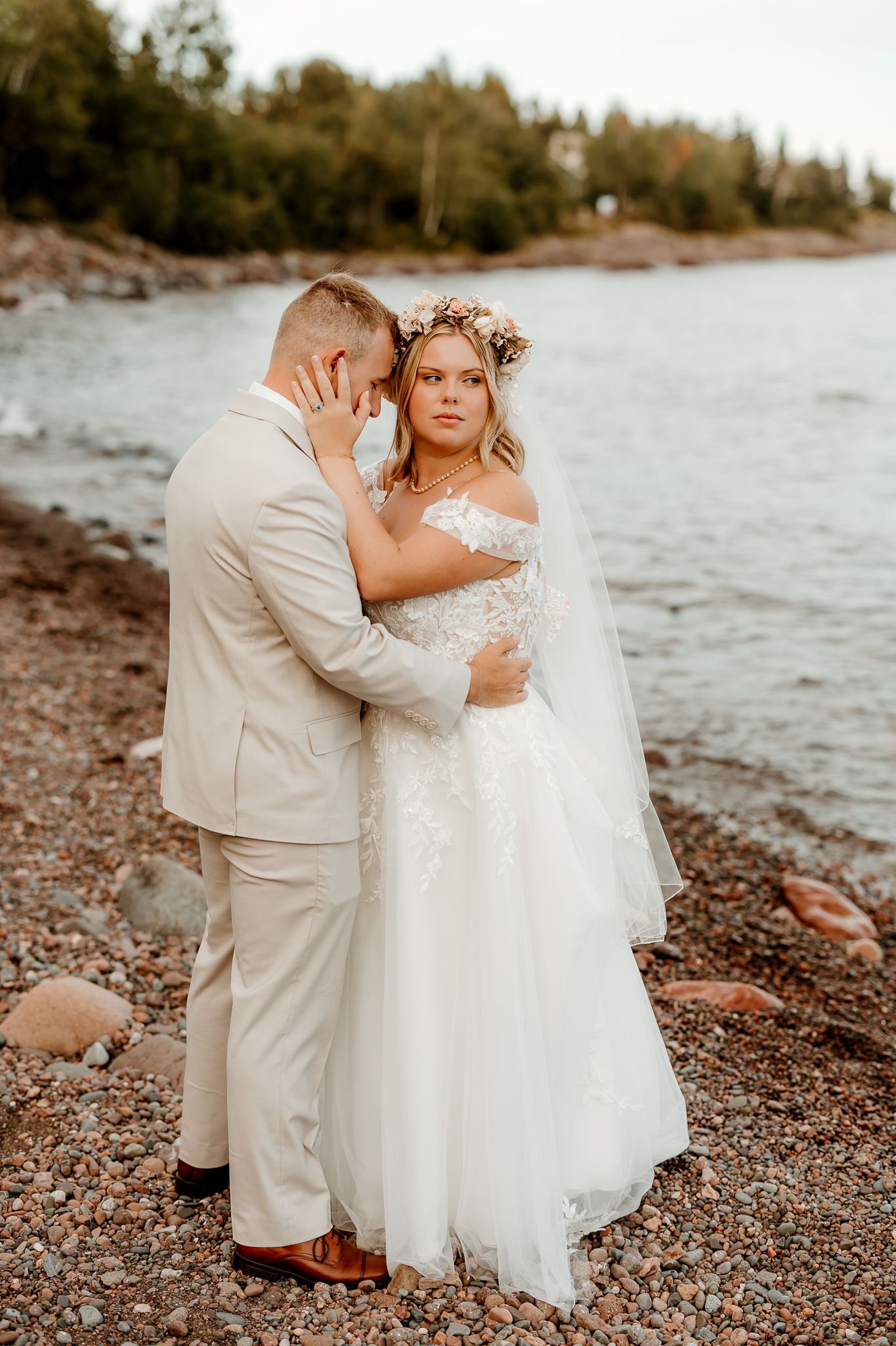 Bride and groom embrace on rocky shoreline, bride wearing floral crown and off-shoulder lace gown, groom in beige suit.