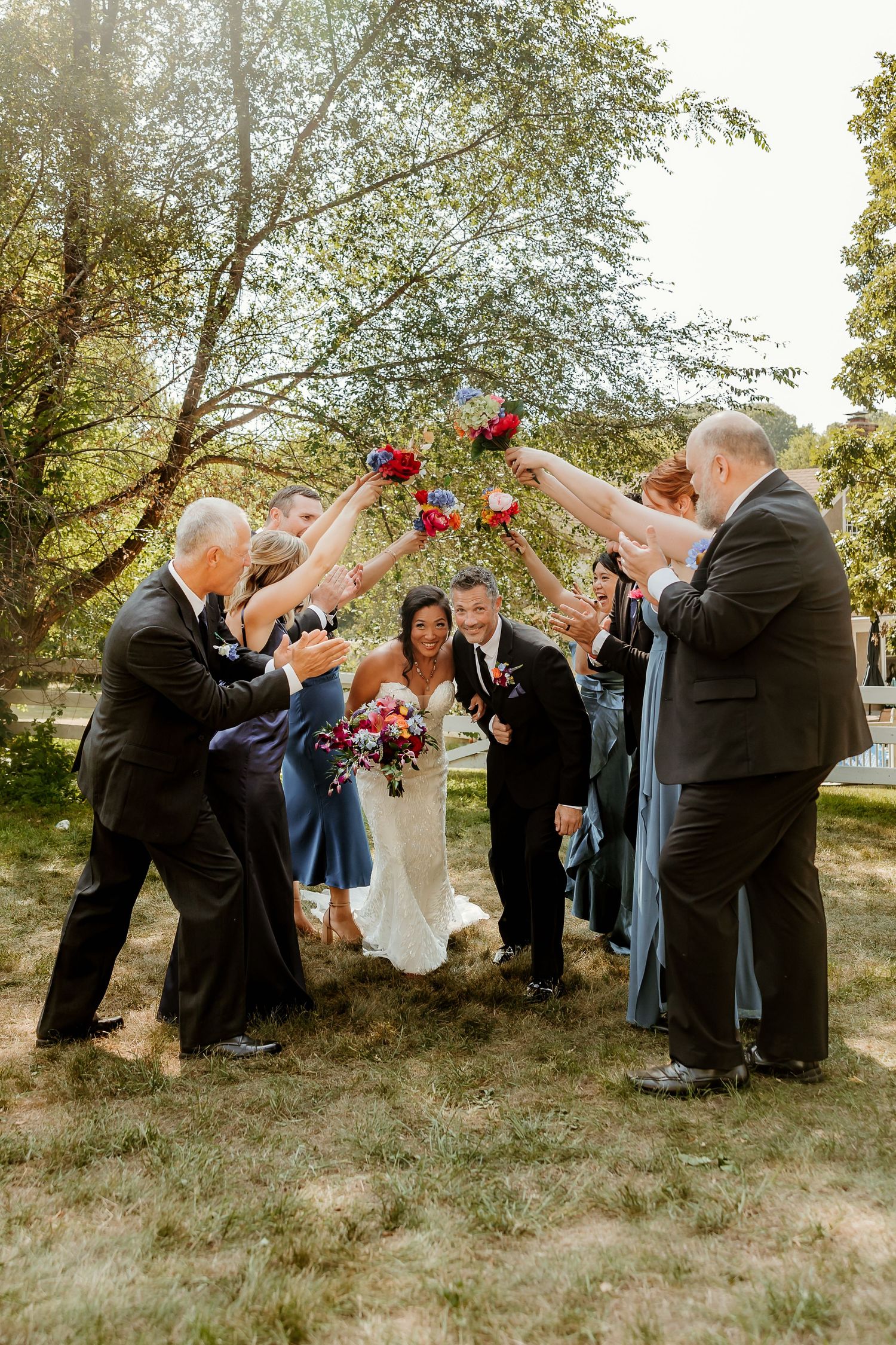 Bride and groom walk through an arch of raised bouquets formed by wedding party guests outdoors.