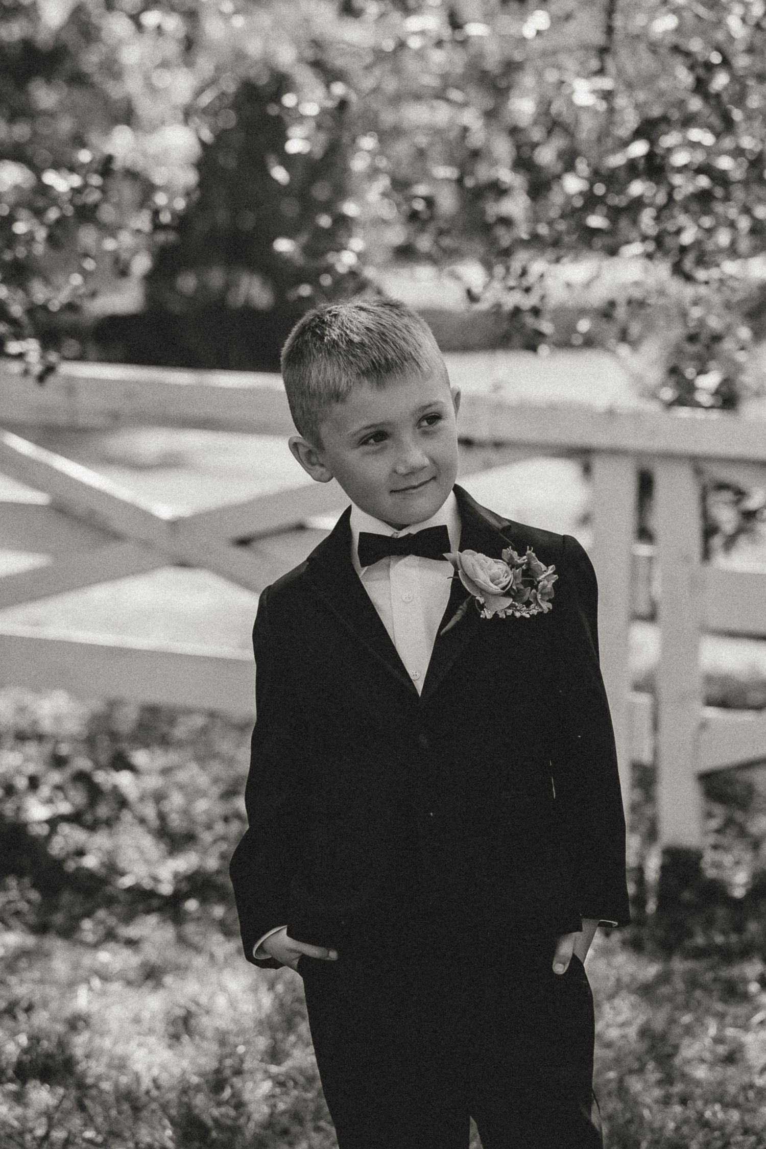 A young ring bearer in a black tuxedo with bow tie and boutonniere poses outdoors in a black and white photo.