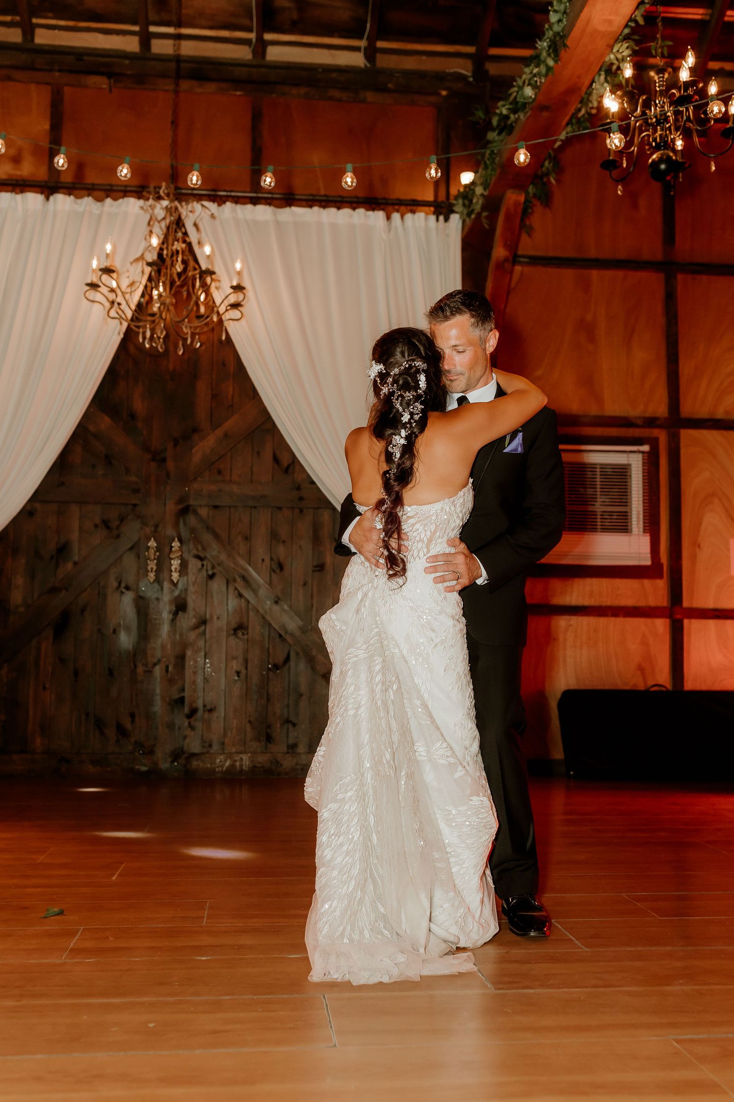 Bride and groom share their first dance in a rustic barn venue adorned with chandeliers and draped white curtains.