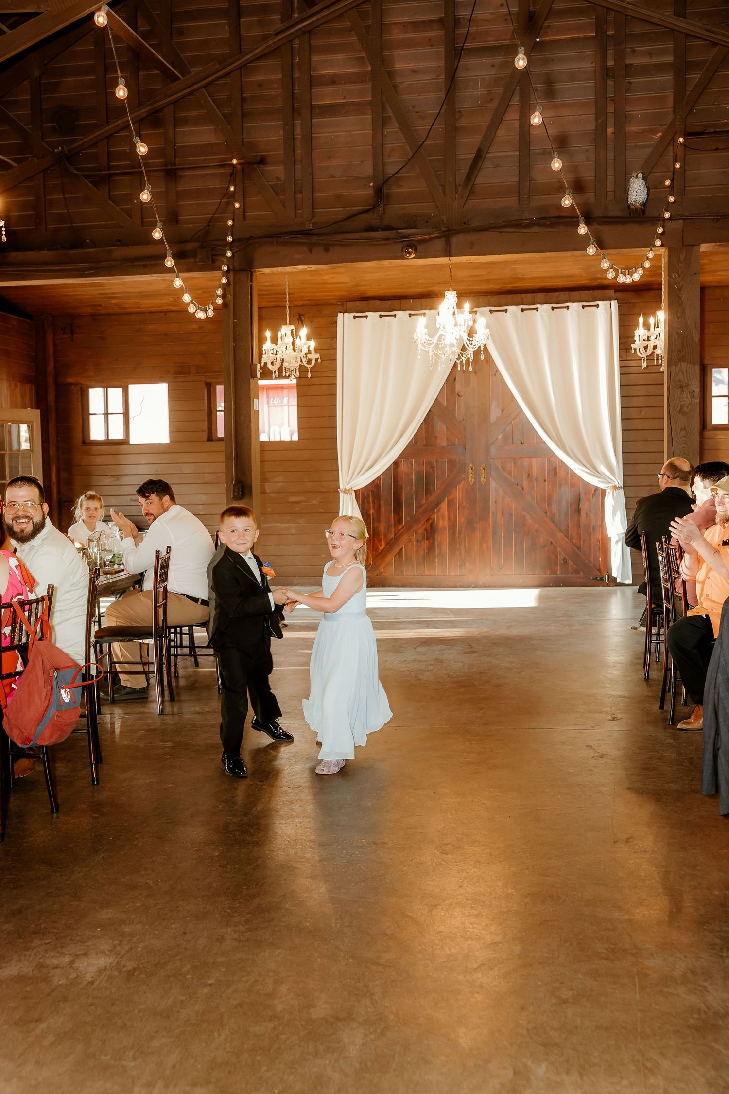 A flower girl in a blue dress and ring bearer in a tuxedo walk down the aisle in a rustic barn wedding venue.