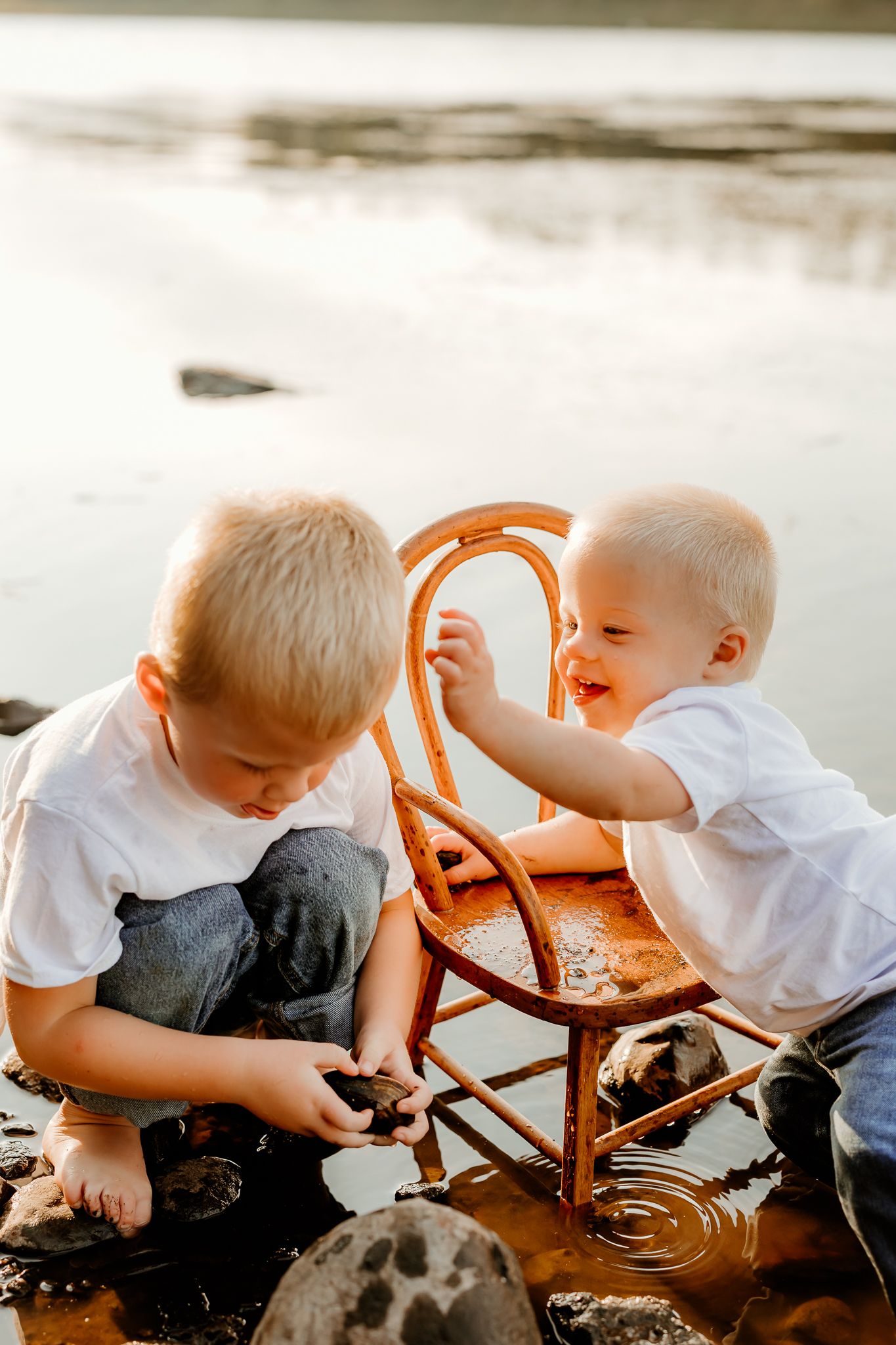 Two young boys in white shirts playing with rocks and a rustic chair in shallow water near a rocky shore.