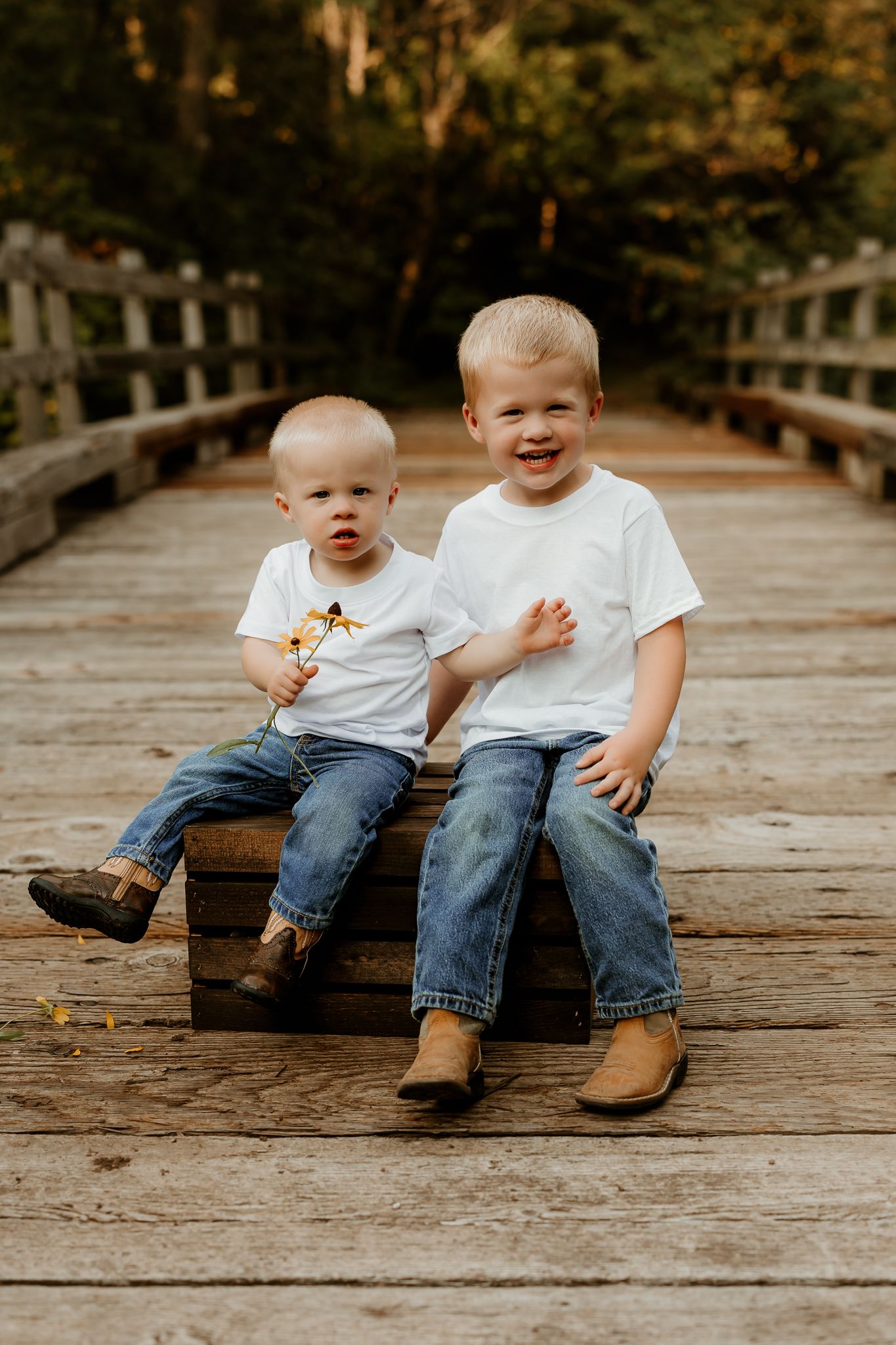Two young blonde boys in white t-shirts and jeans sit together on a wooden bridge surrounded by autumn trees.