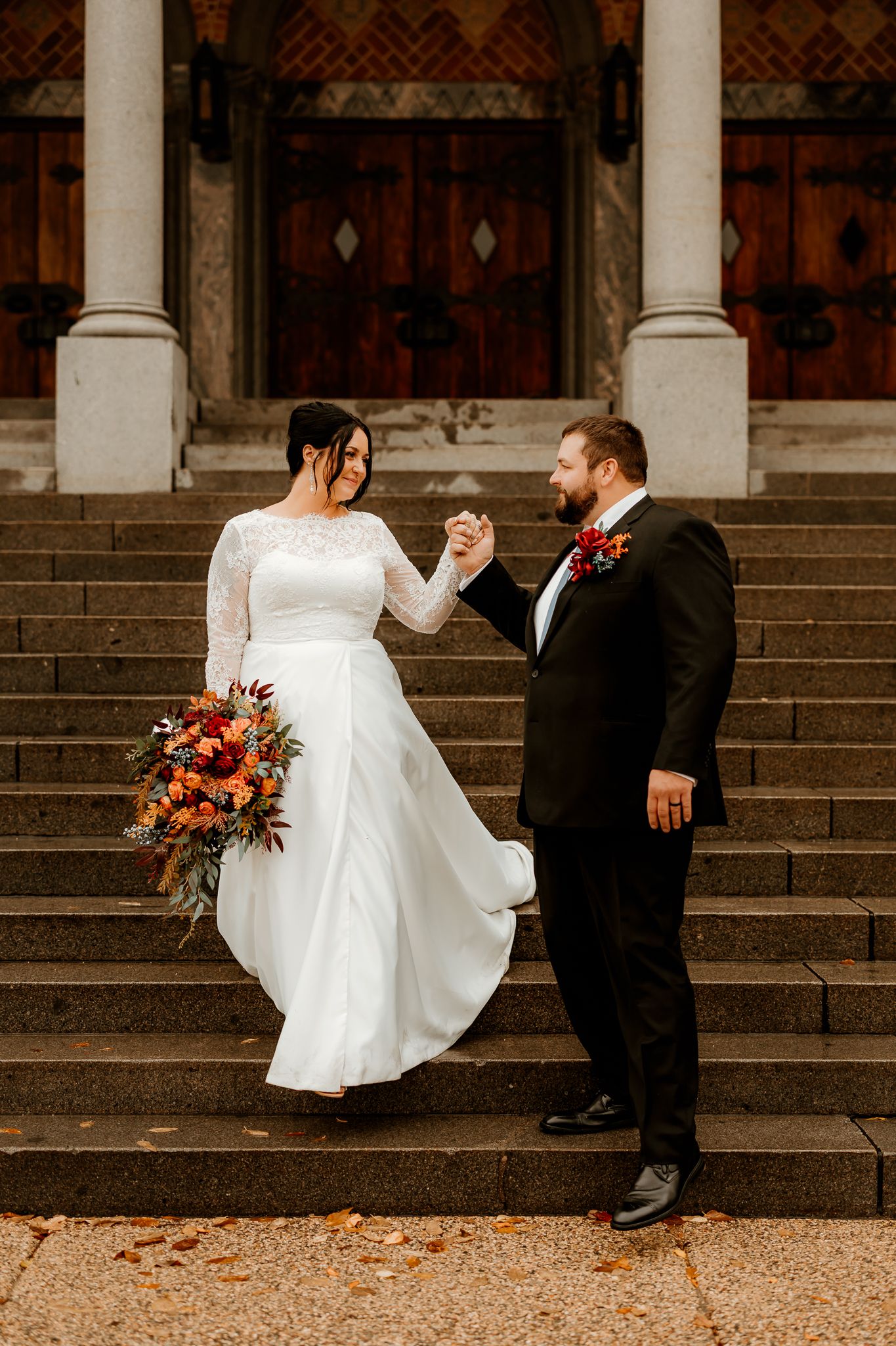 Bride in a white lace gown holding an autumn bouquet walks hand in hand with a groom in a dark suit down church steps.