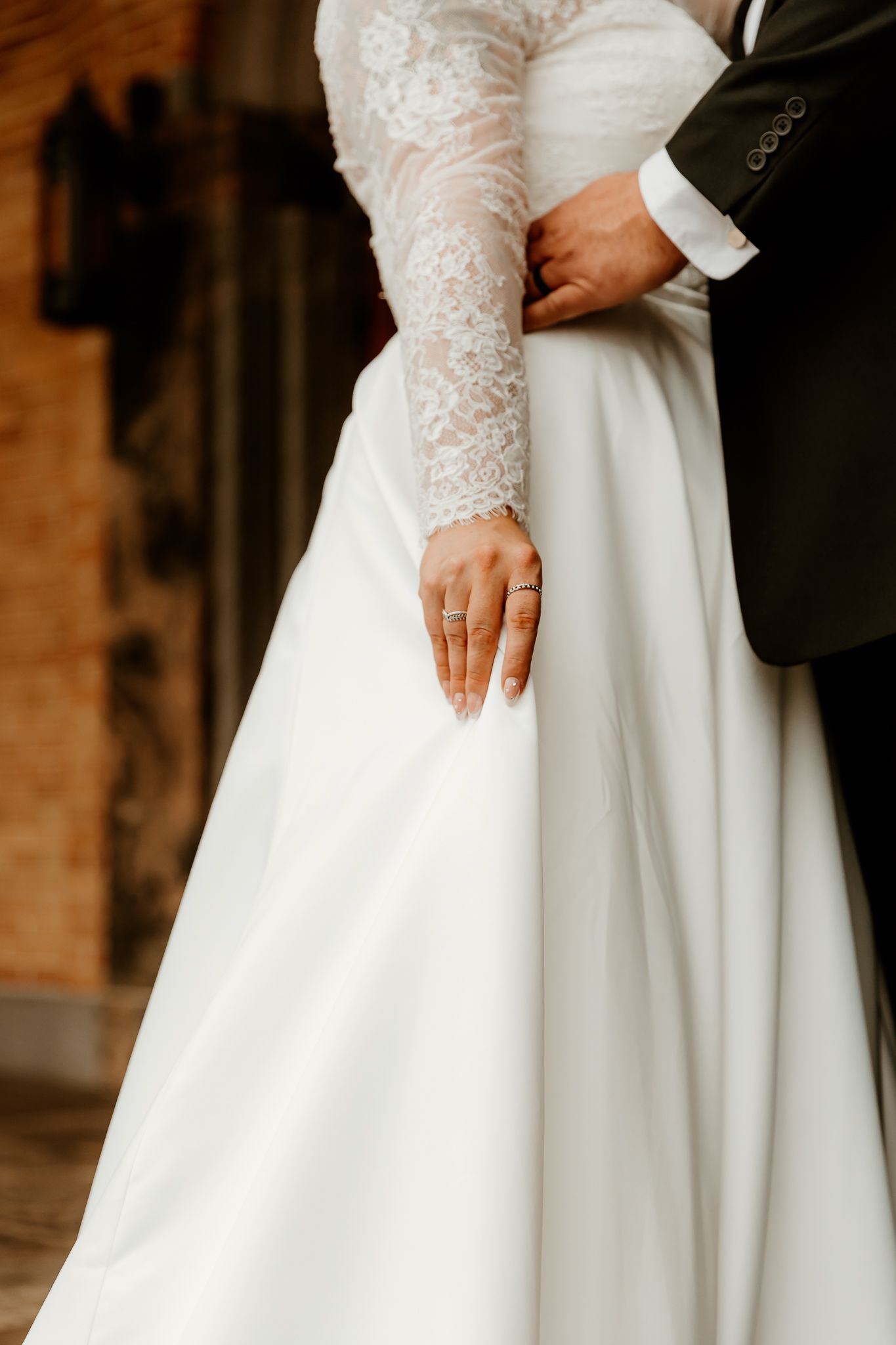 Bride in a white satin wedding gown with lace sleeves and a ring on her hand embraced at the waist by a groom in a dark suit.