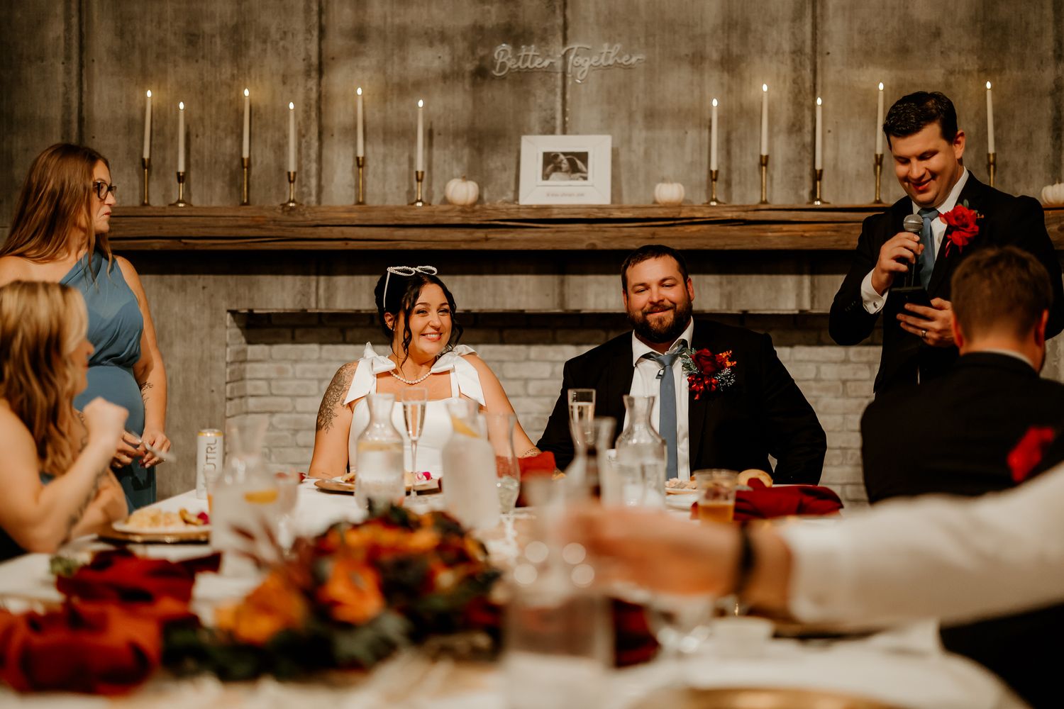 Bride and groom sit at a candlelit reception table as a guest in a suit gives a toast, with autumn floral centerpieces in the foreground.