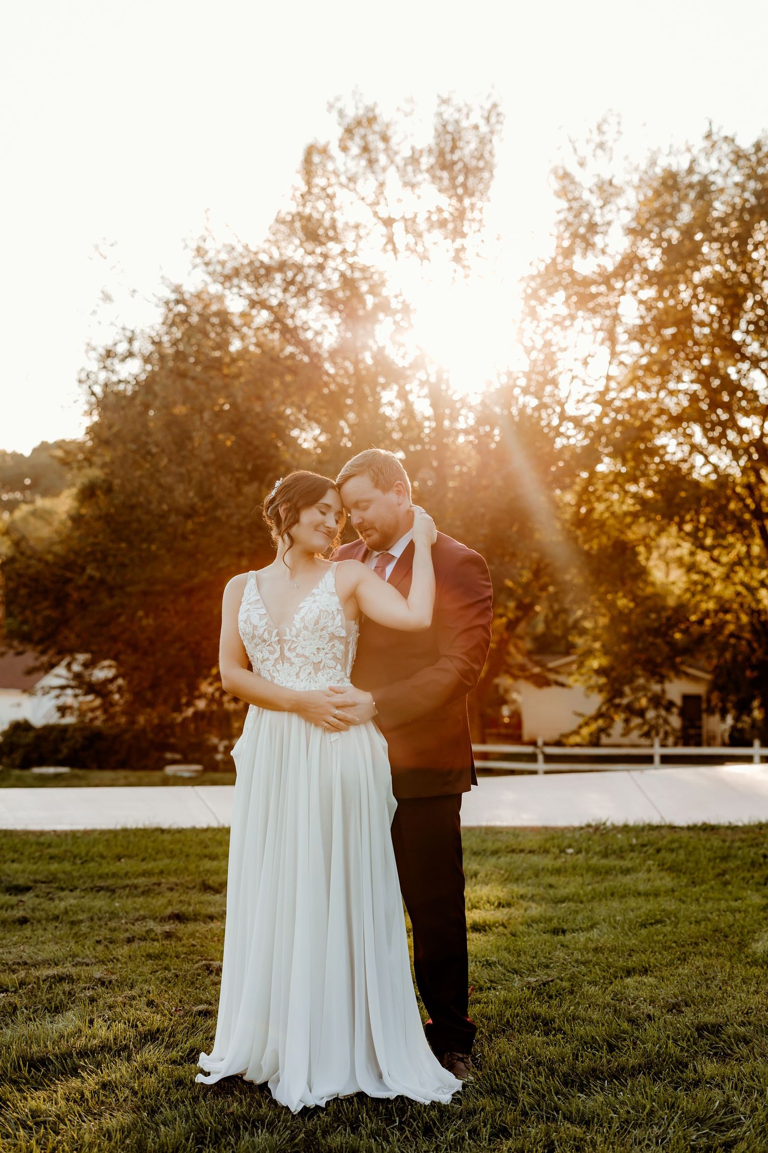 Bride in a flowing lace gown and groom in a burgundy suit embrace on a grassy lawn at golden hour with sunlight streaming through trees.