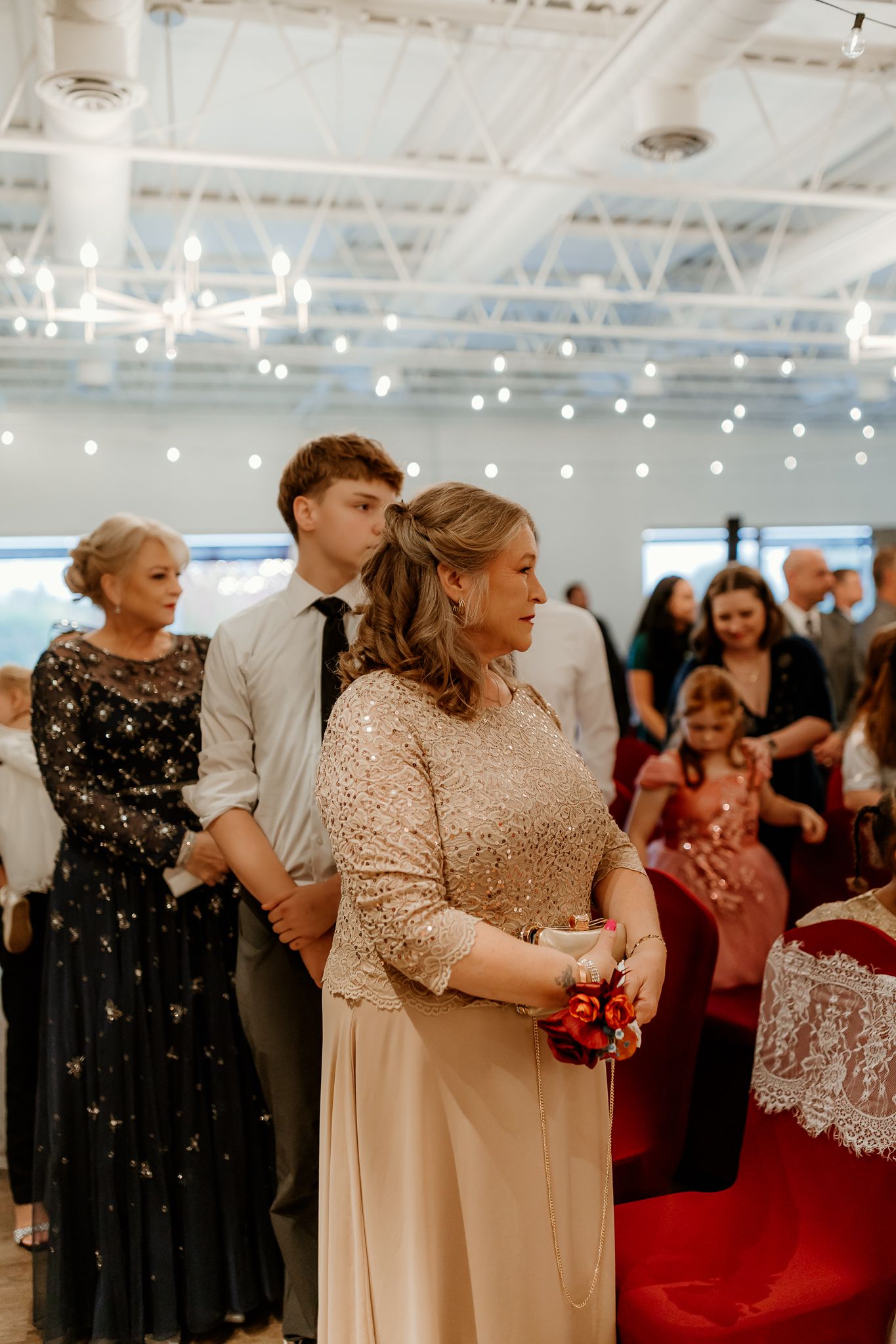 Wedding guests stand during a ceremony in a bright event hall, with a woman in a champagne gown holding a red bouquet in the foreground.