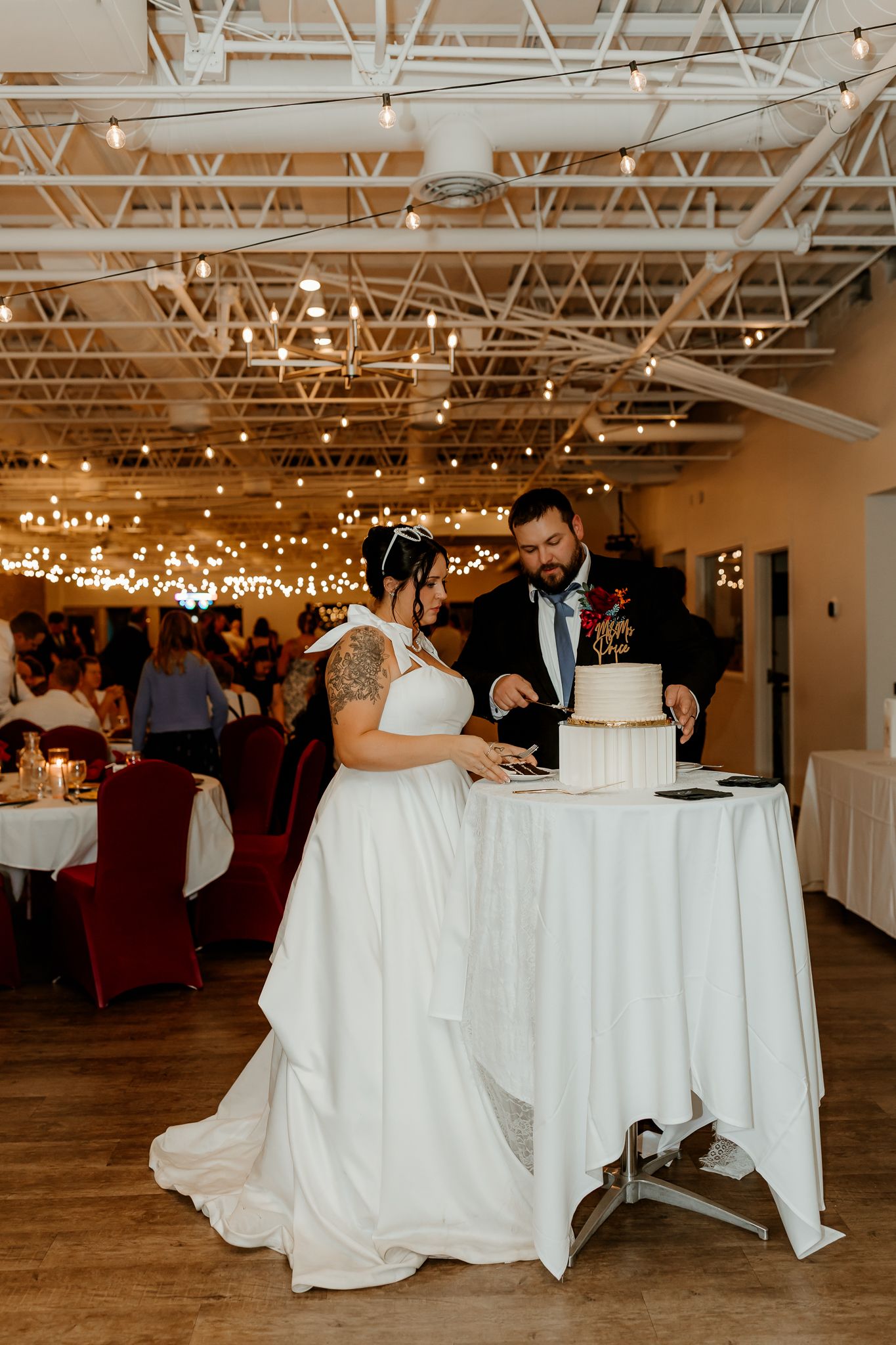 Bride in a white gown and groom in a dark suit cut their white tiered wedding cake at a reception hall with string lights.