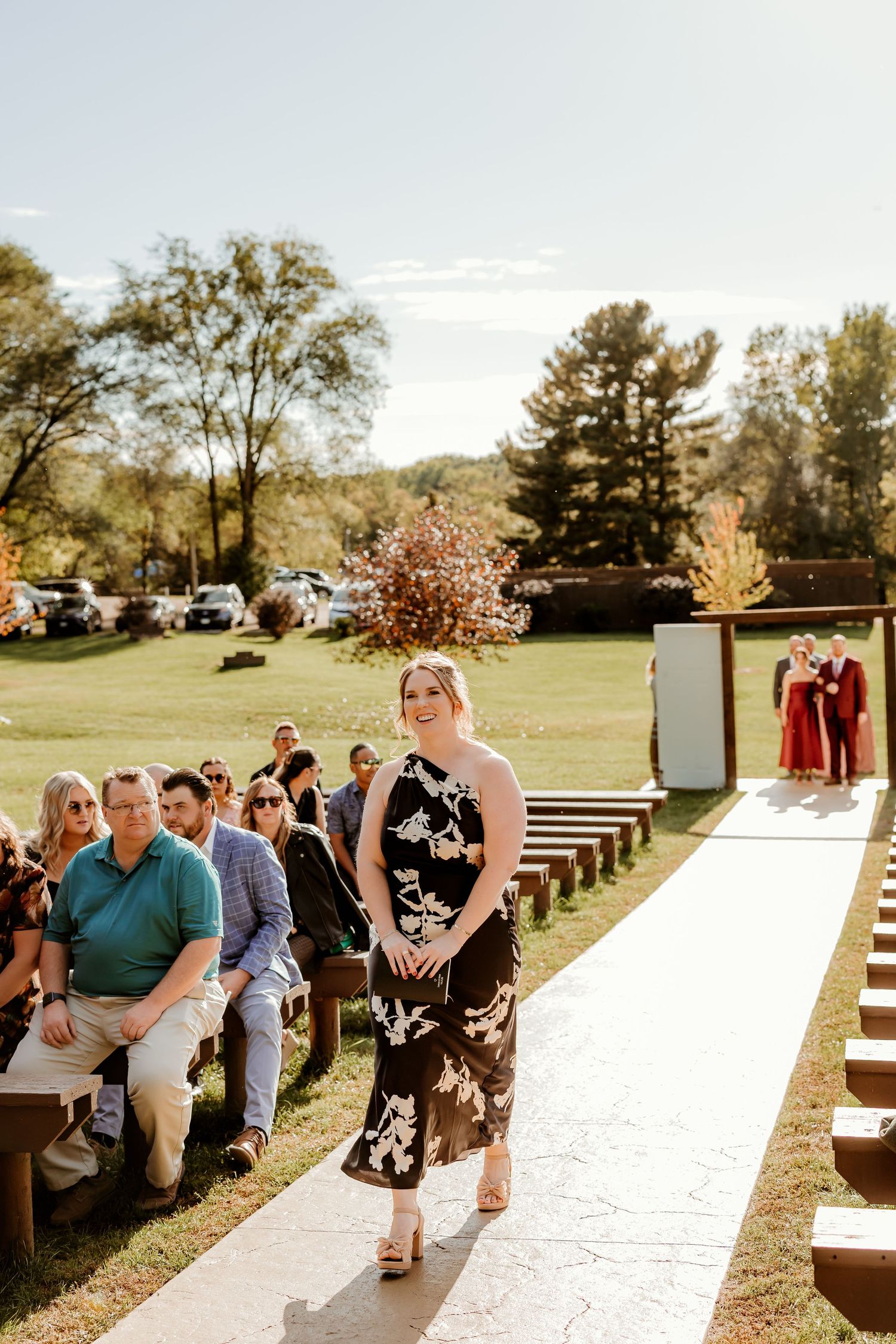Woman in a black floral dress walks down the aisle of an outdoor wedding ceremony as seated guests watch under autumn trees.