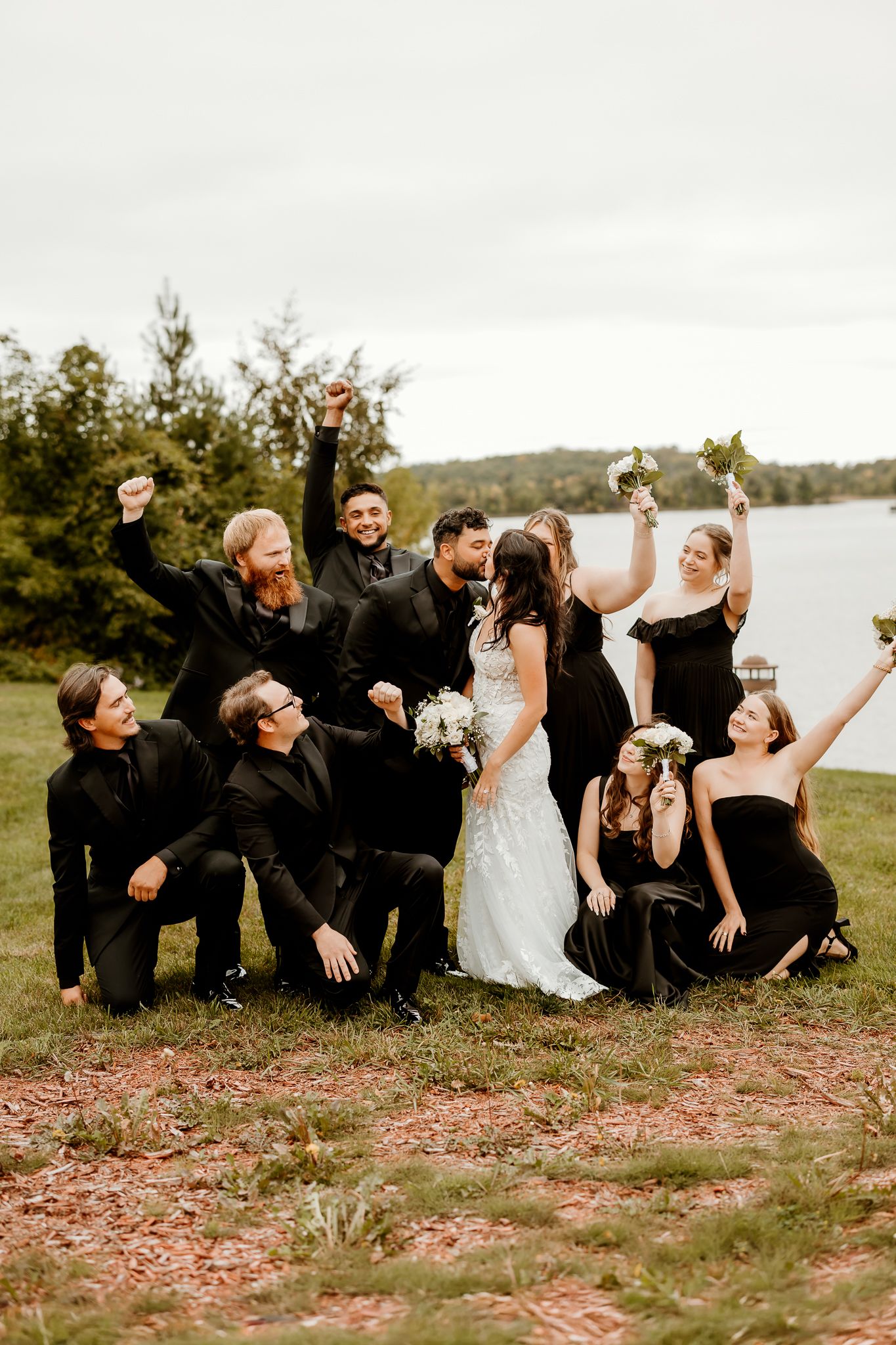 Bride in a white lace gown laughs with her wedding party in black attire, cheering and raising bouquets on a grassy lakeside lawn.