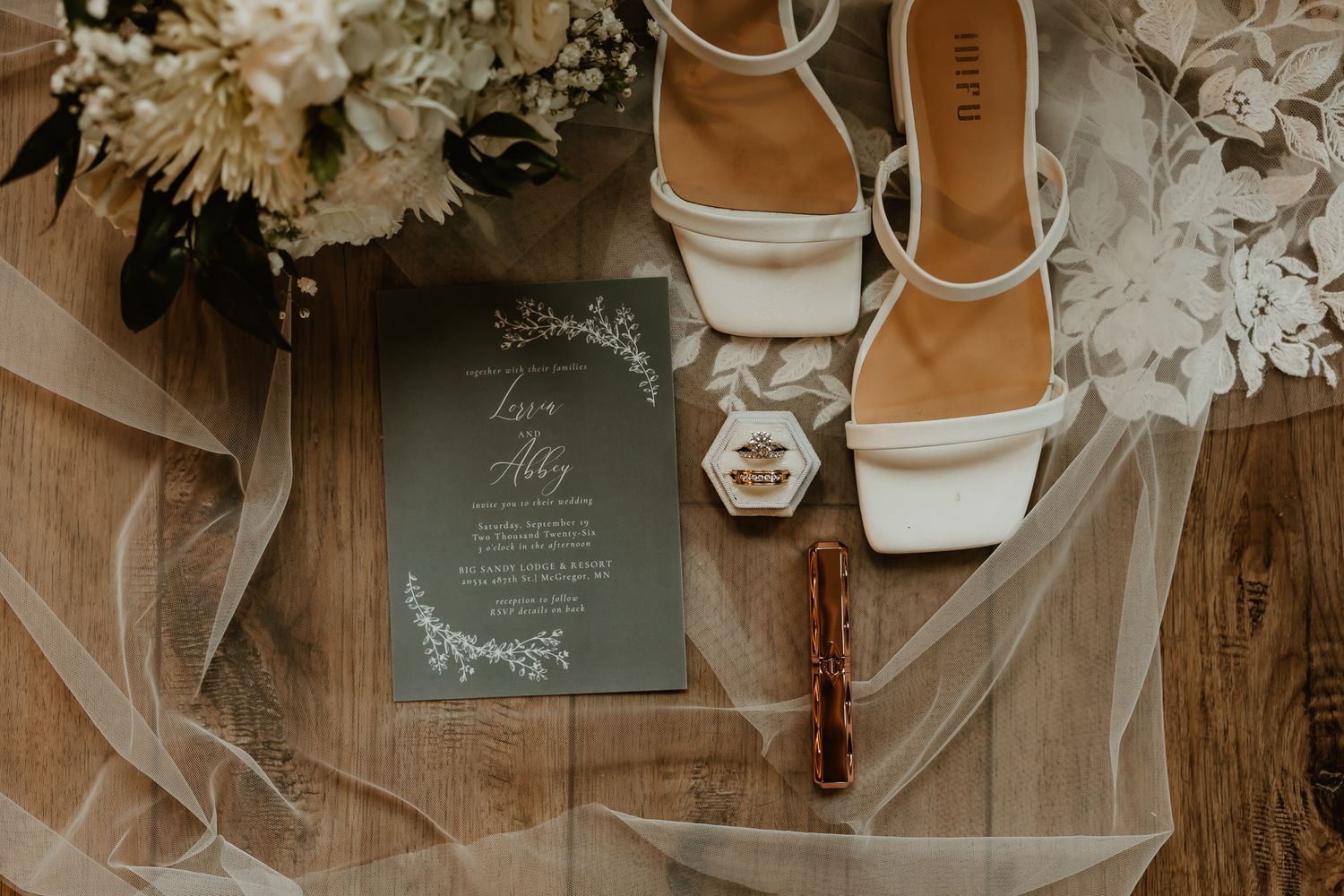 Wedding flat lay with white strappy heels, gray invitation, ring box, lipstick, tulle veil and floral bouquet on a rustic wooden surface.