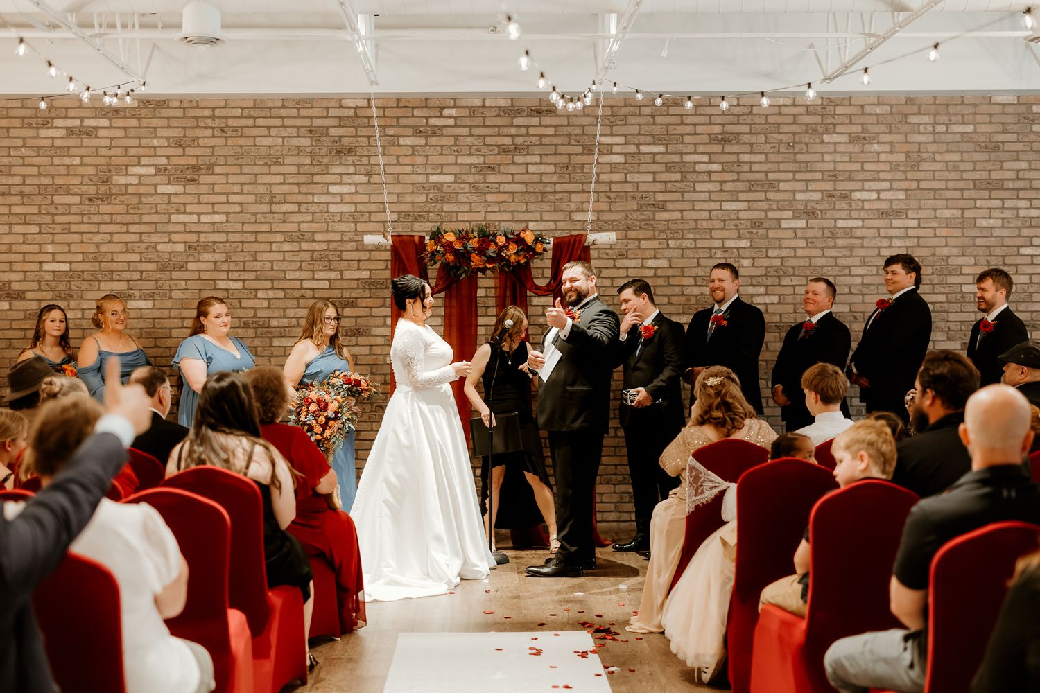 Bride in a white gown and groom in a dark suit exchange vows at an indoor brick-walled wedding ceremony as guests in red chairs look on.