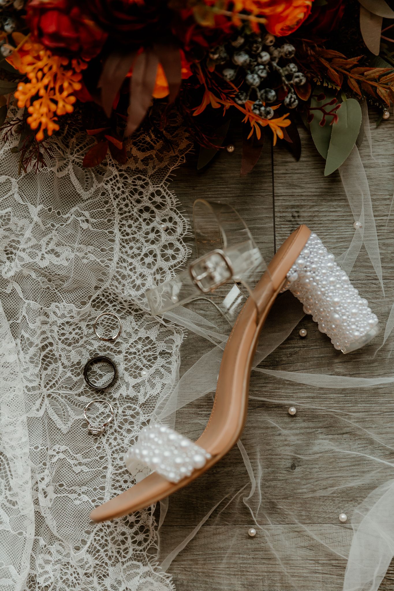 Wedding flat lay with pearl-embellished nude heels, rings on lace, crystal glasses and an autumn orange bouquet on a rustic wood floor.