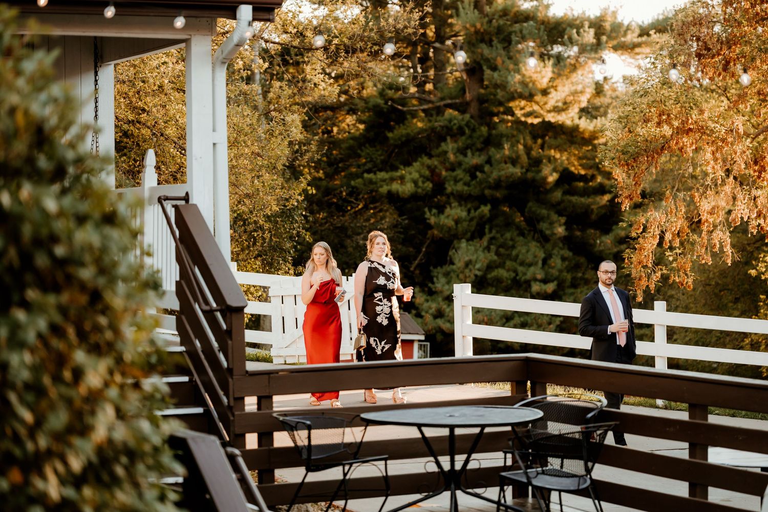 Two women in red and floral dresses chat on a wooden deck while a man in a suit stands nearby, surrounded by golden autumn trees.