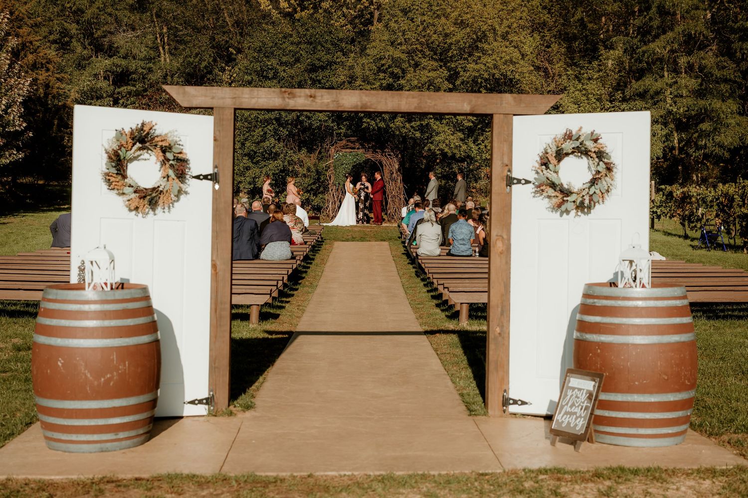 Outdoor rustic wedding ceremony with guests seated on wooden benches along an aisle framed by white doors with wreaths and whiskey barrels.