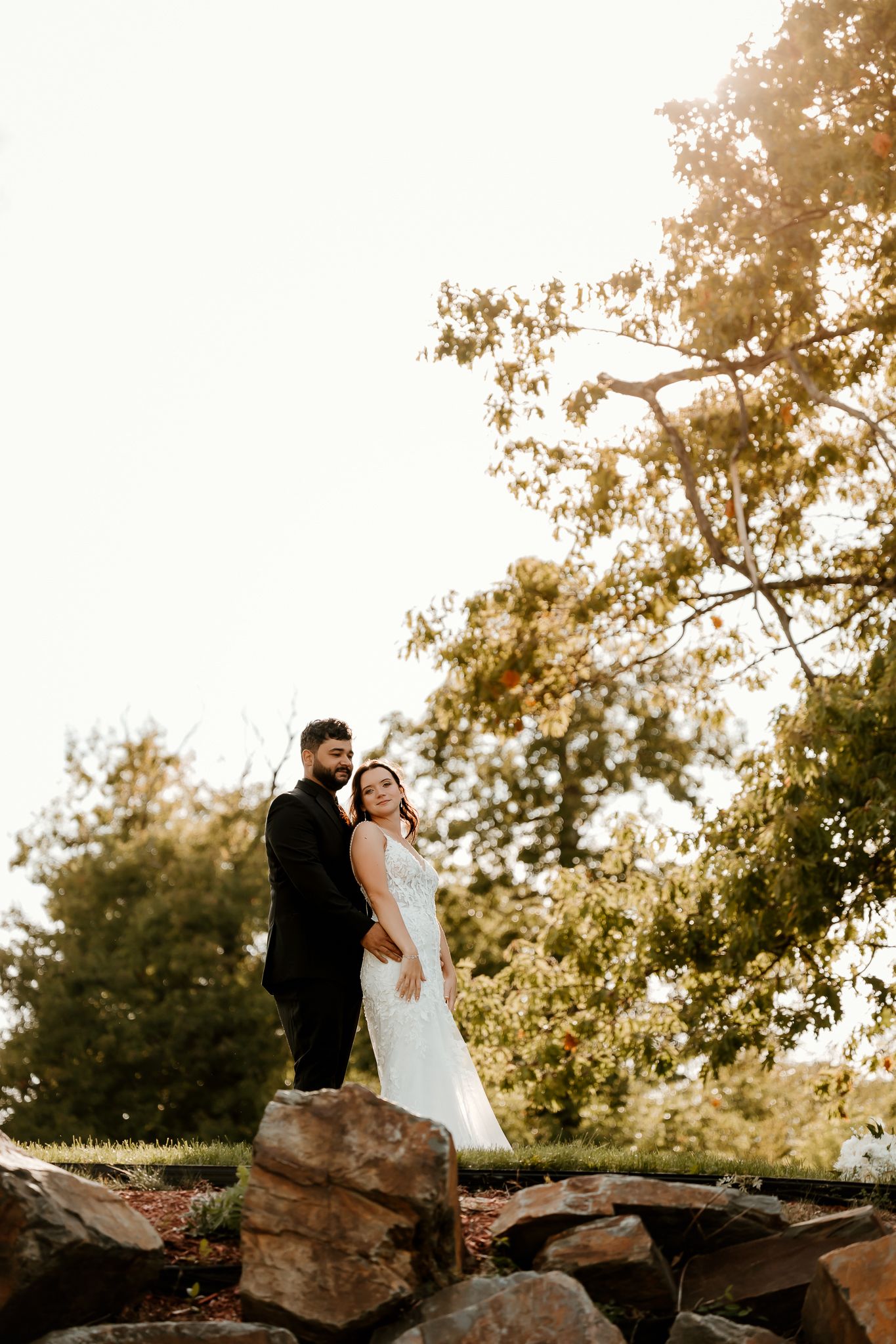 Bride in a white lace gown embraces her groom in a black suit on a rocky hillside surrounded by sunlit autumn trees.