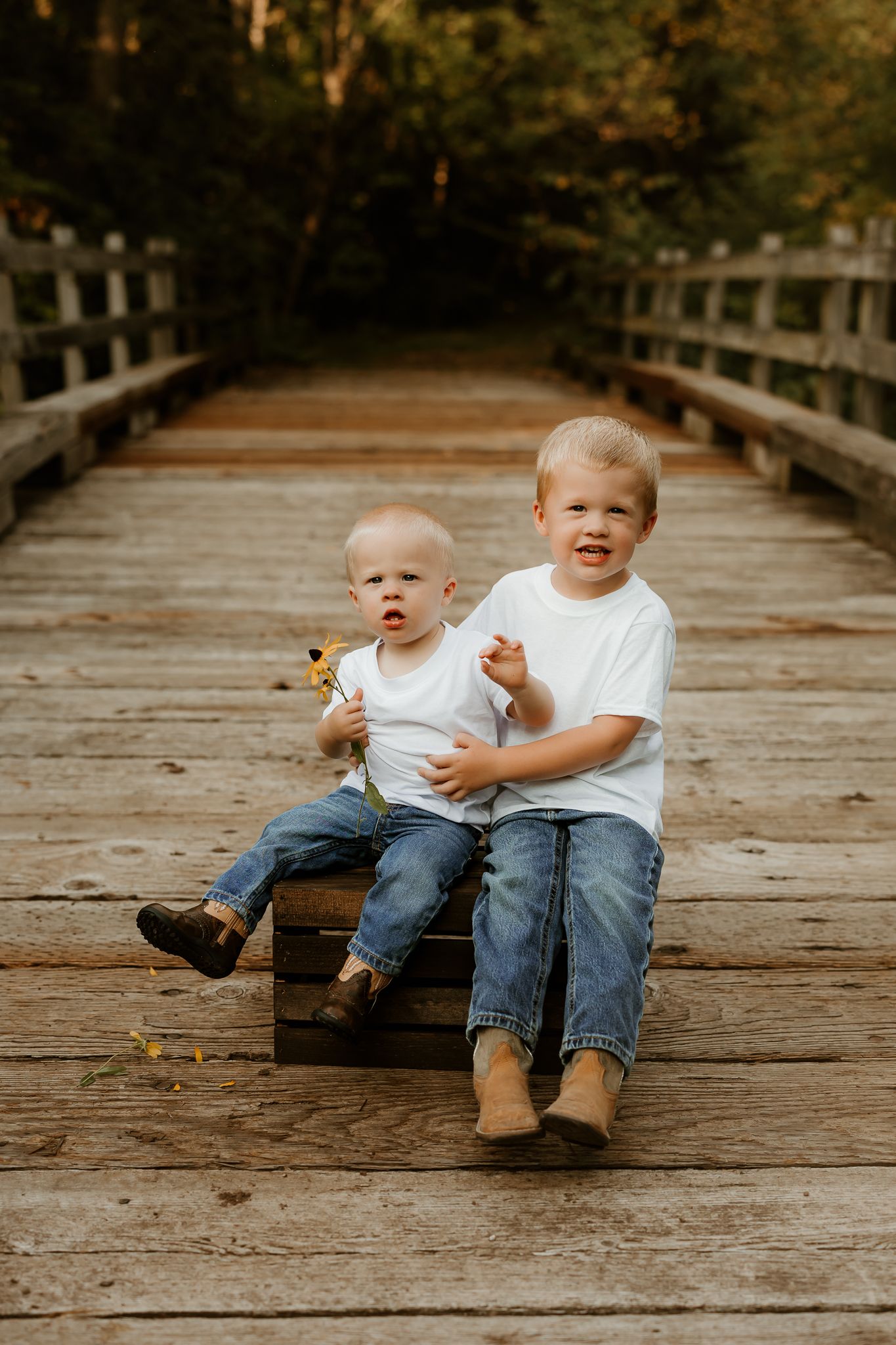 Two young boys in white shirts and jeans sitting on a wooden crate on a bridge surrounded by trees.