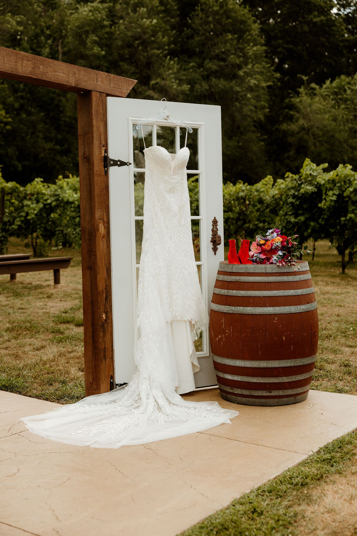 White lace wedding dress hanging on a rustic door frame beside a wine barrel with floral decor in a vineyard setting.