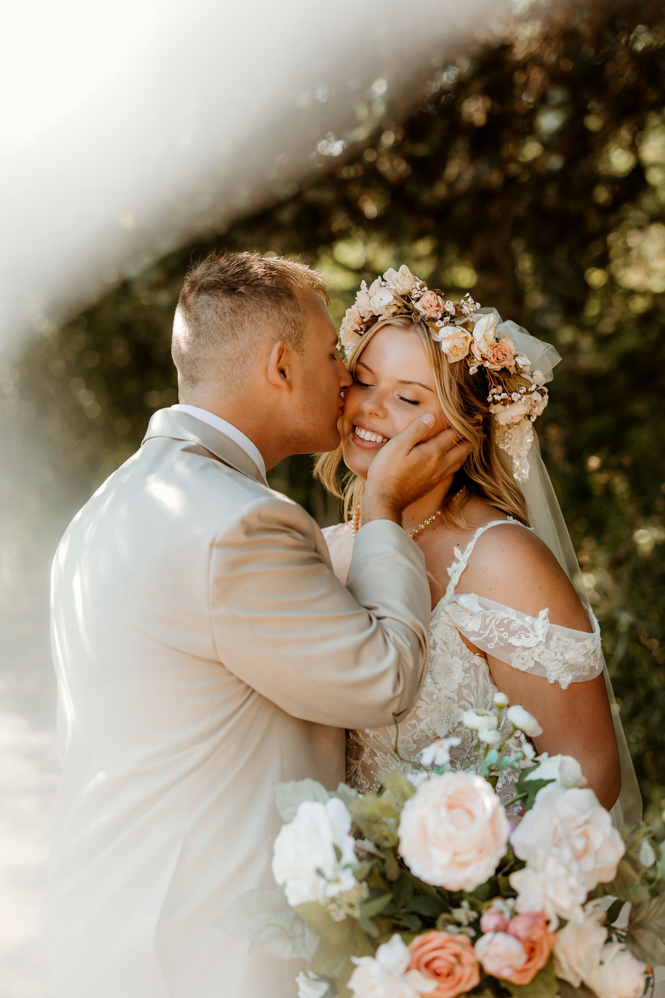 Bride smiling as groom kisses her cheek, she wears a floral crown and holds a pastel bouquet.