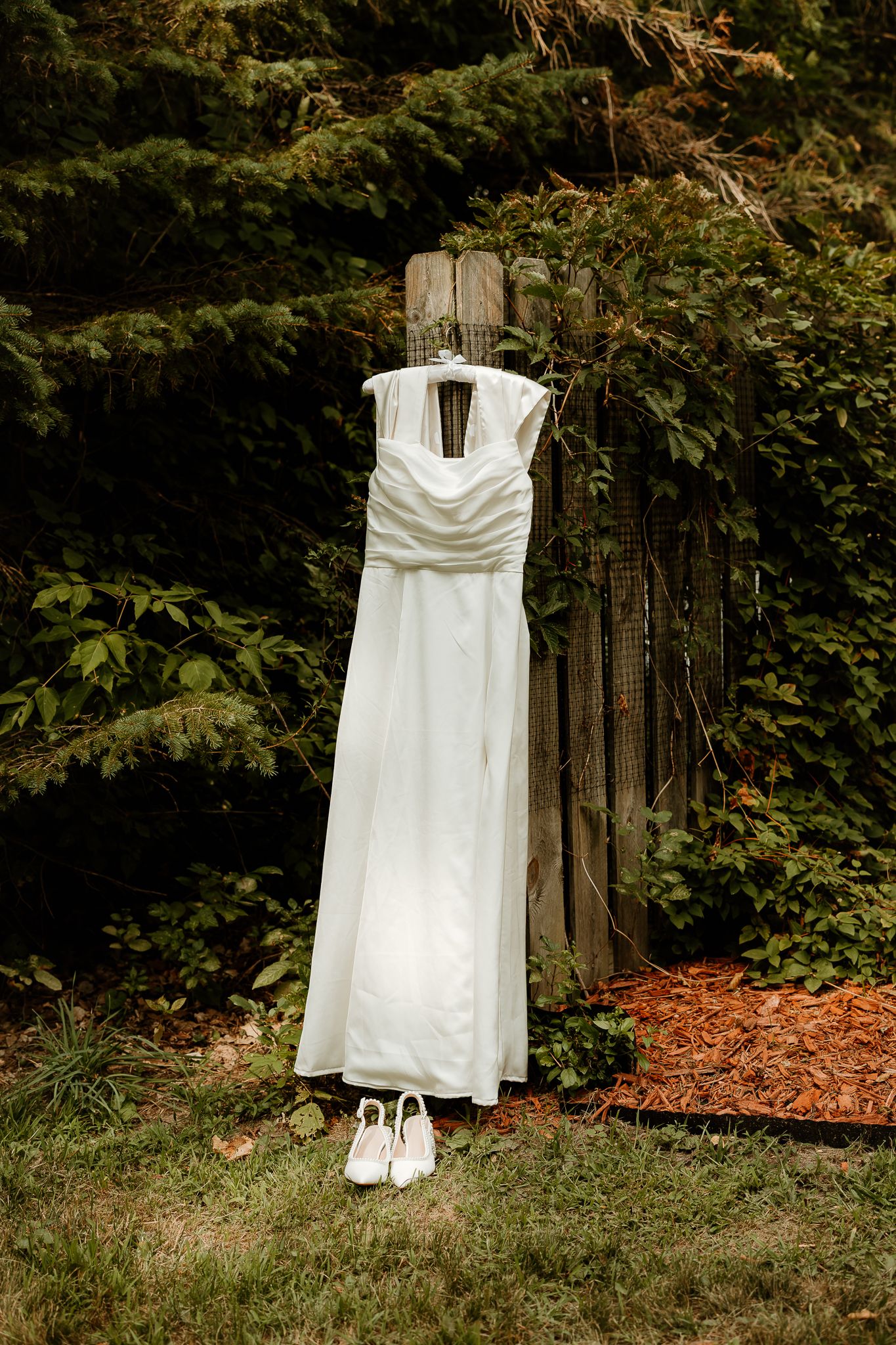 White wedding dress hanging outdoors on wooden post with matching bridal heels on grass below.