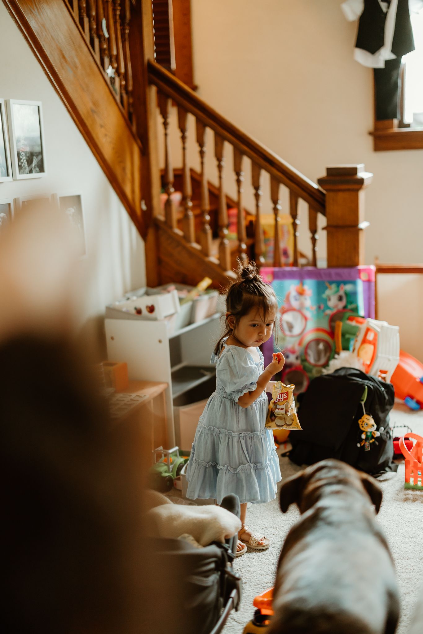 A young girl in a blue dress plays with toys near a wooden staircase while a dog rests nearby in a cozy home.