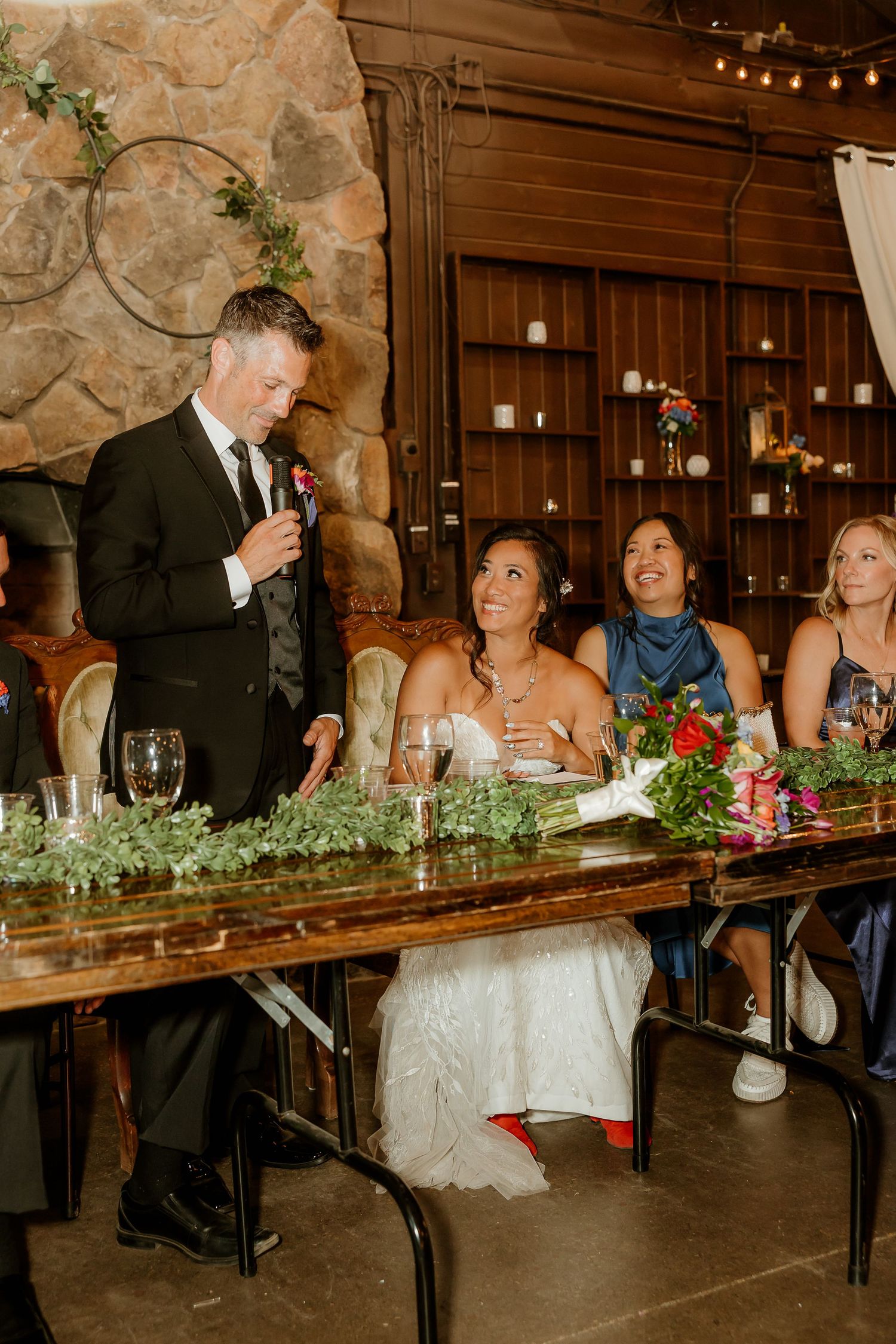 Groom in black tux gives wedding toast as bride and bridesmaids smile at rustic wood reception venue.