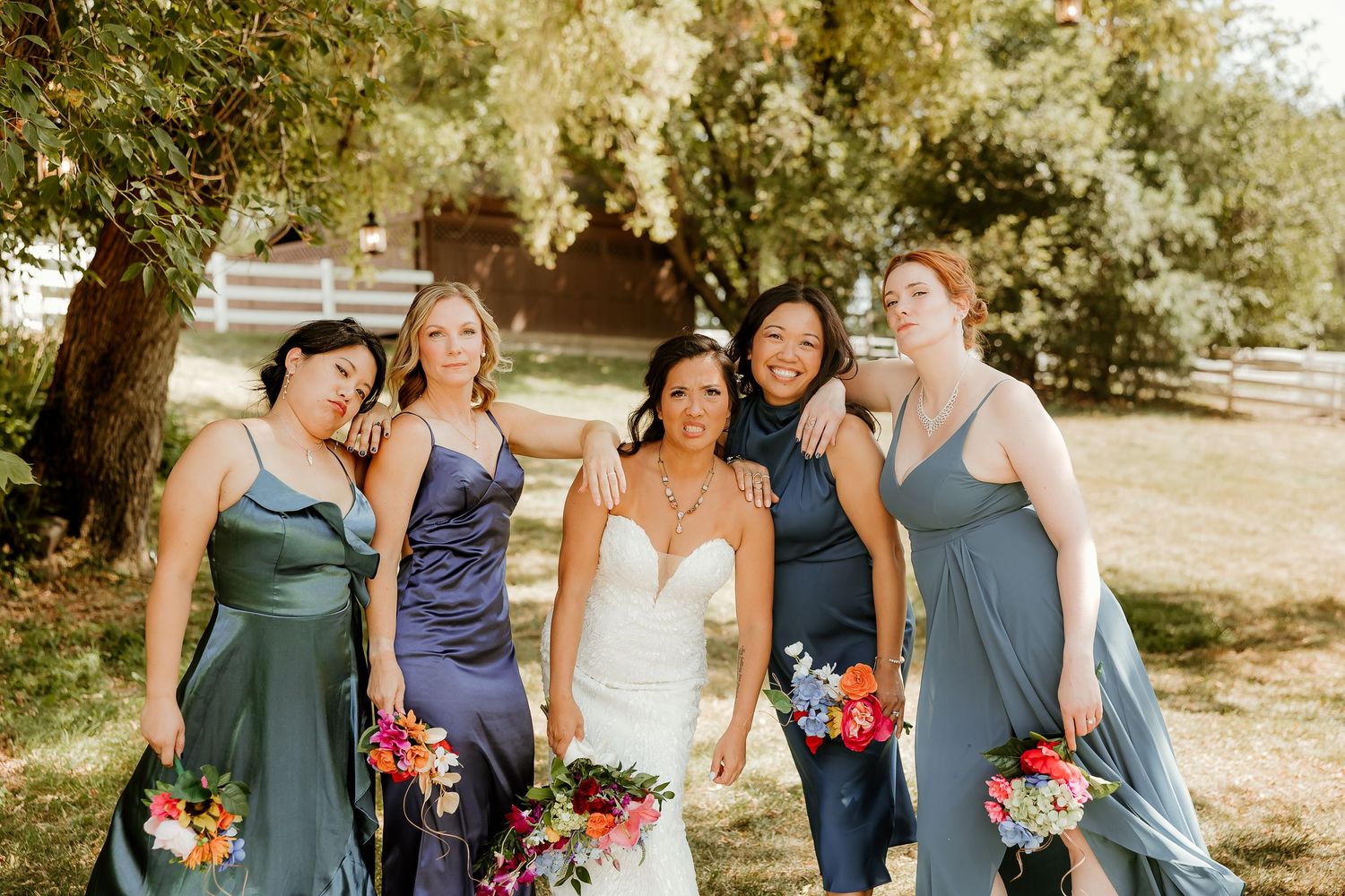 Bride in white gown poses with four bridesmaids in blue and green dresses holding colorful bouquets outdoors.