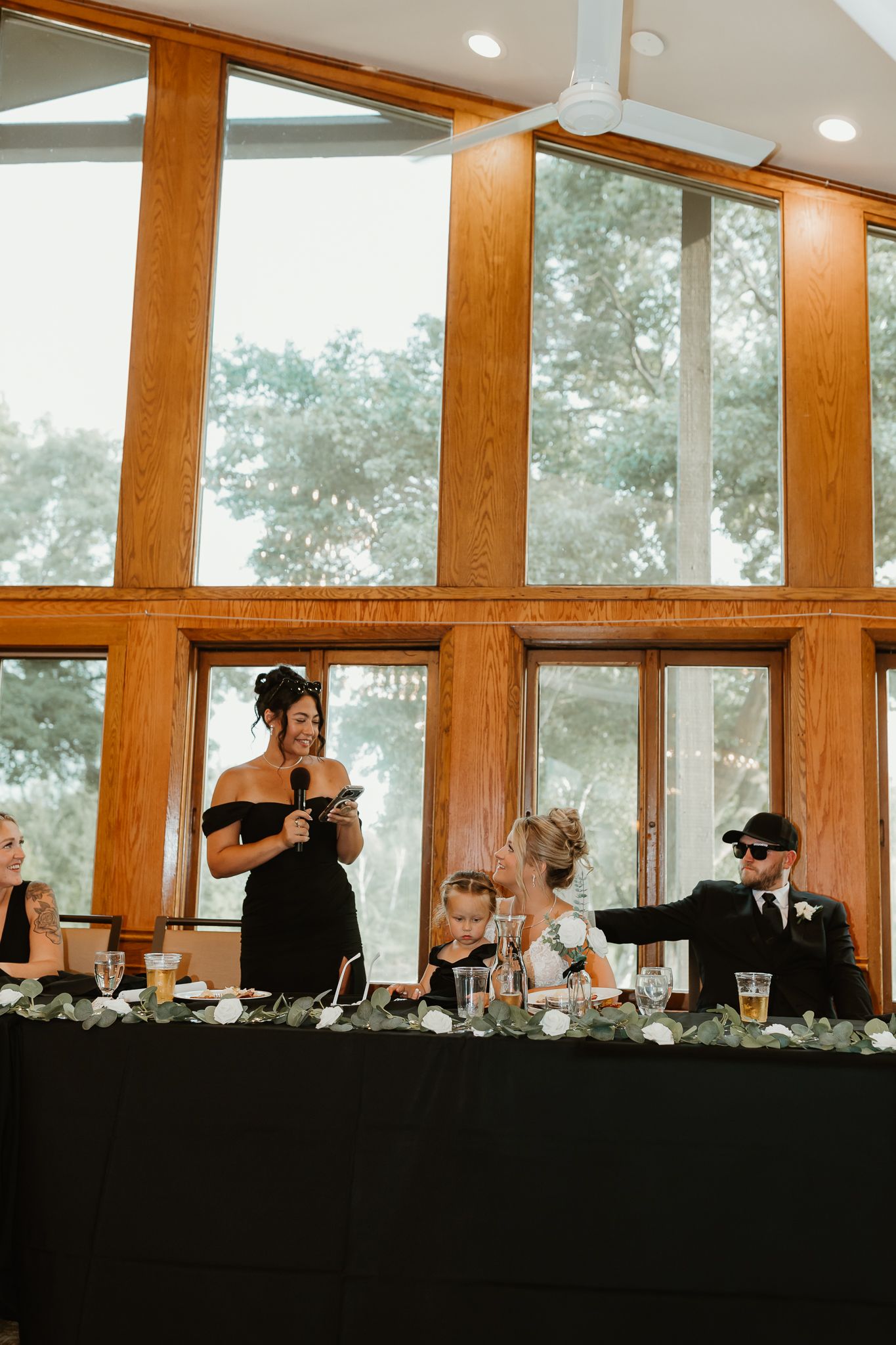 A bridesmaid in a black dress gives a toast at a wedding reception with large wooden-framed windows in the background.