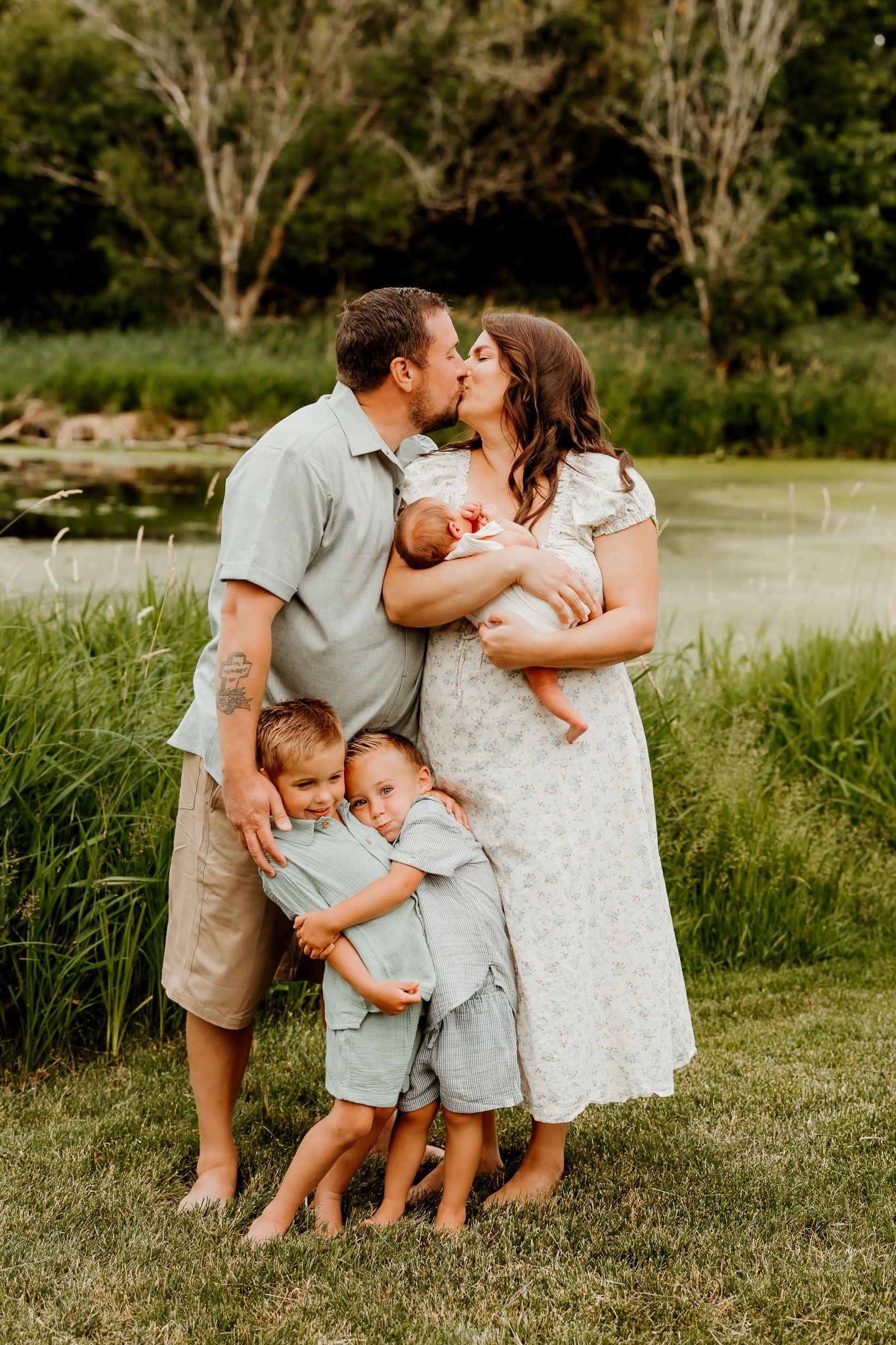 Family of five poses outdoors by a pond, parents kissing while holding newborn, two young boys standing in front.