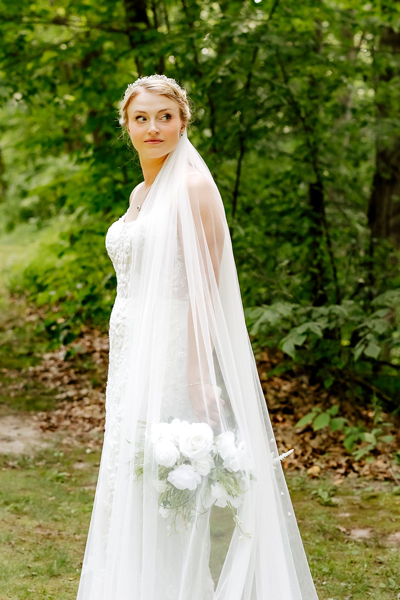 Elegant bride in white lace gown and long veil holds white bouquet outdoors in lush green forest setting.