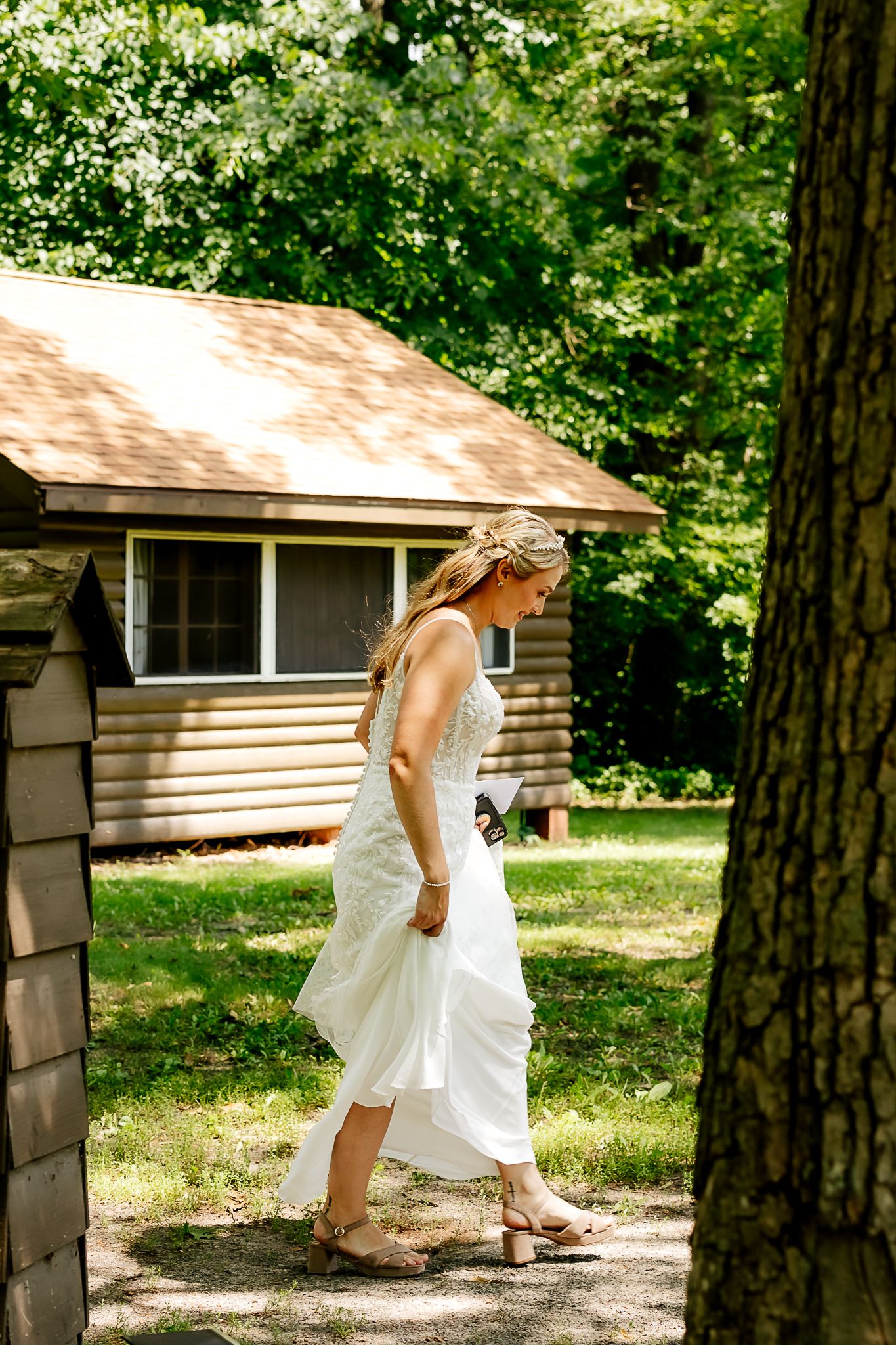 Bride in white gown walks through wooded camp setting with log cabin backdrop on her wedding day.