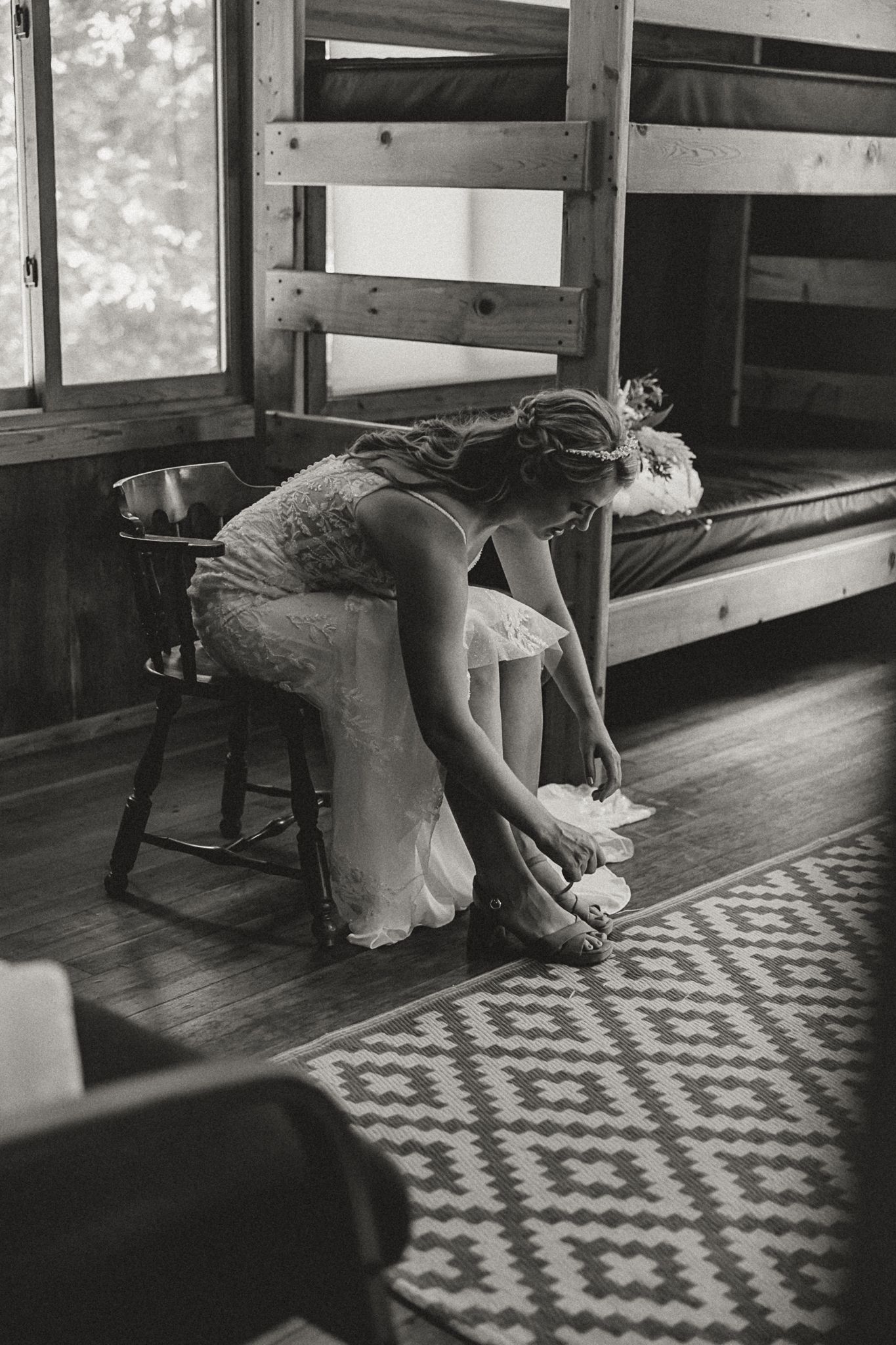 Bride in lace wedding dress putting on shoes while seated in rustic cabin with bunk beds, black and white photo.