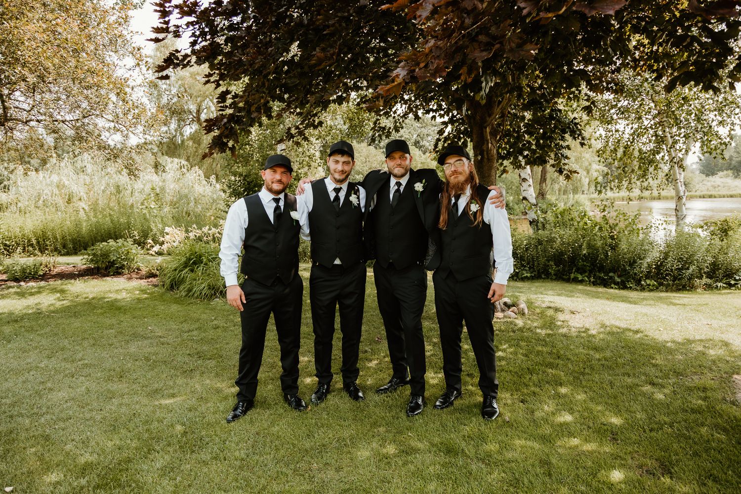 Four groomsmen in black suits and vests pose together outdoors under a tree at a wedding celebration.