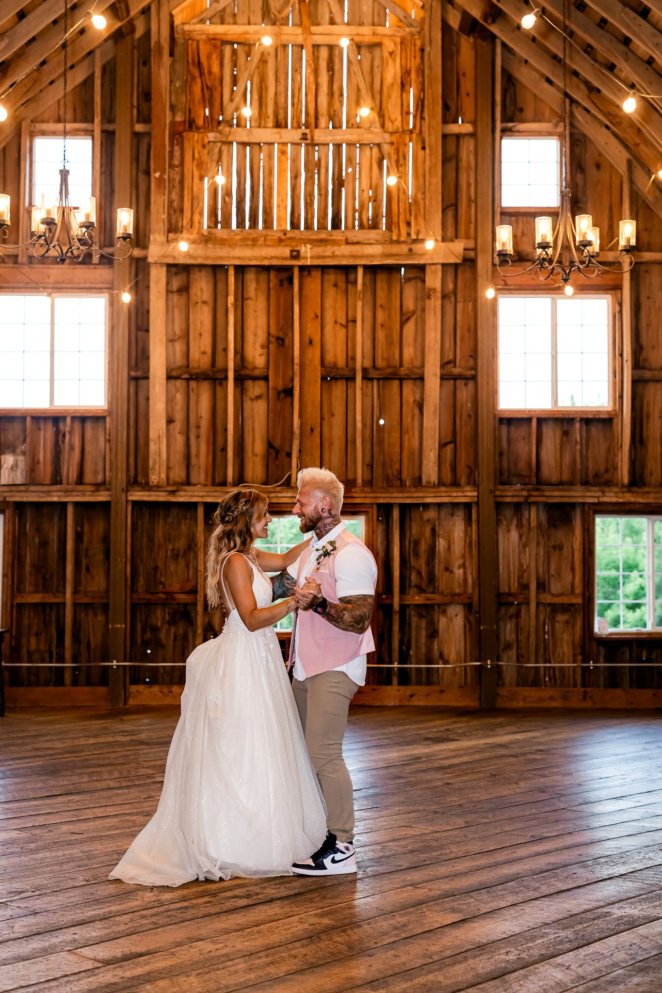 Bride and groom share first dance in rustic barn wedding venue with warm string lights and wooden walls.