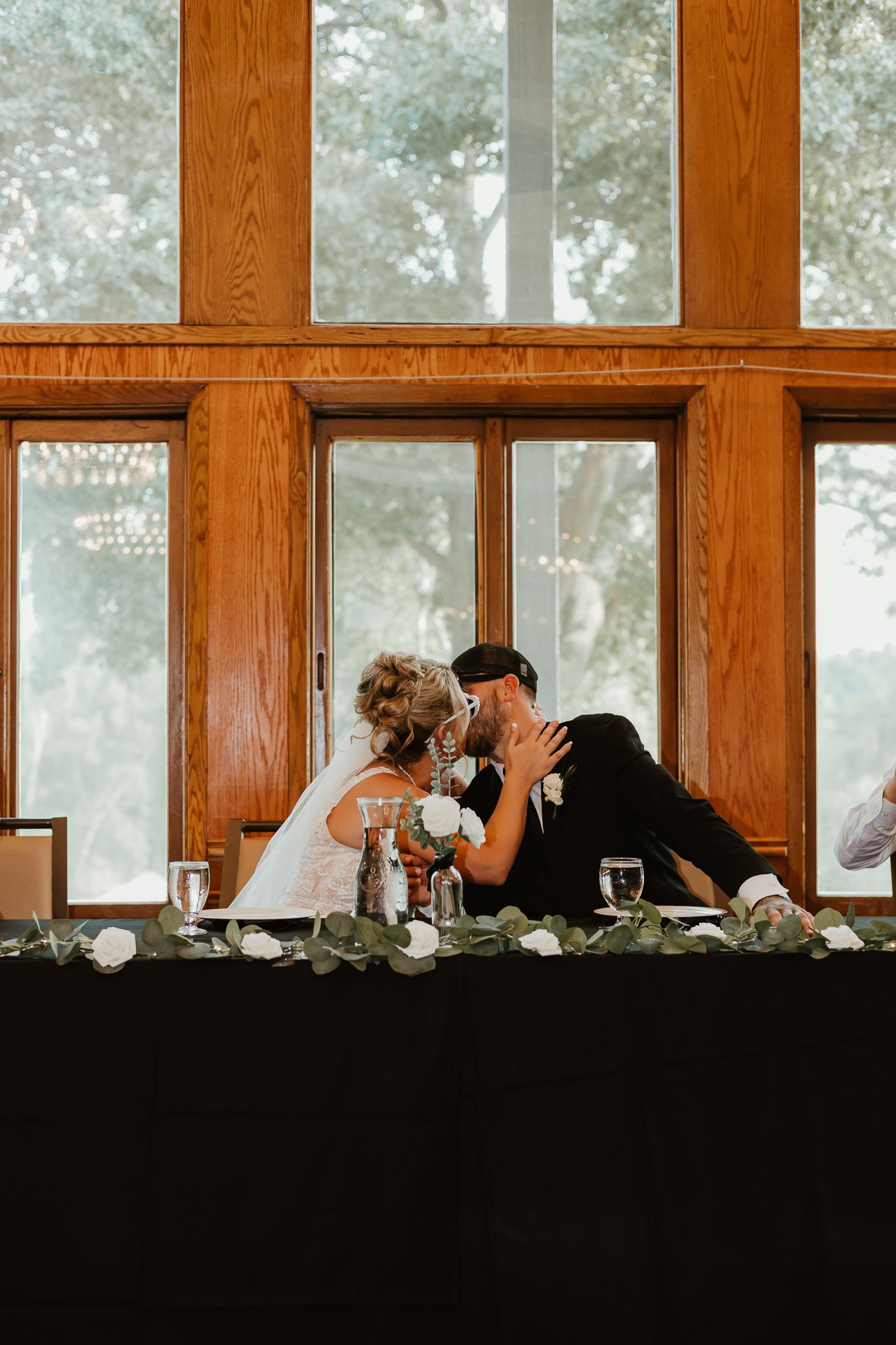 Bride and groom sharing a kiss at their wedding reception head table, decorated with greenery garland and white roses.