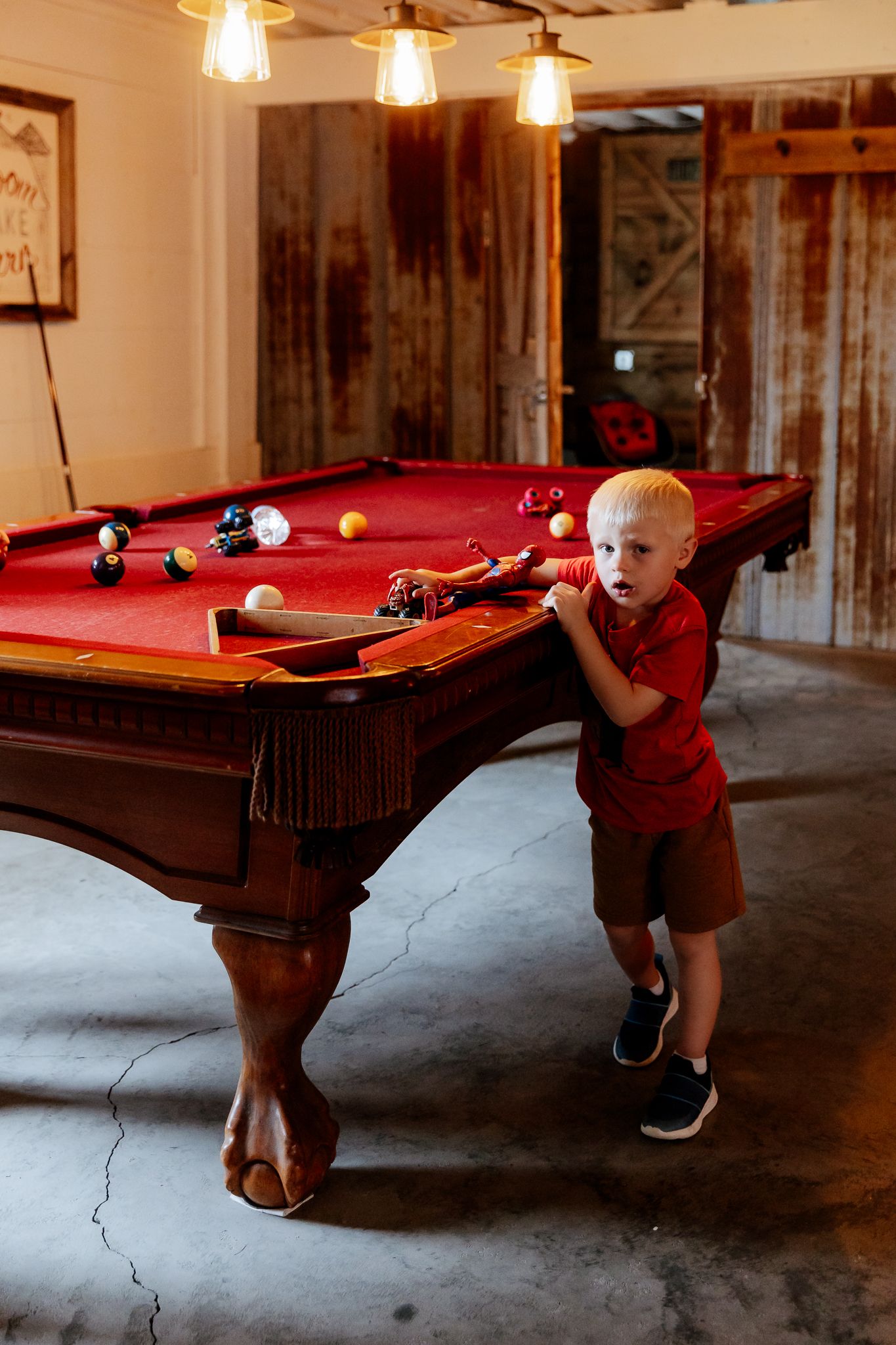 Young blonde boy in red shirt playing pool on a red billiard table in a rustic game room.