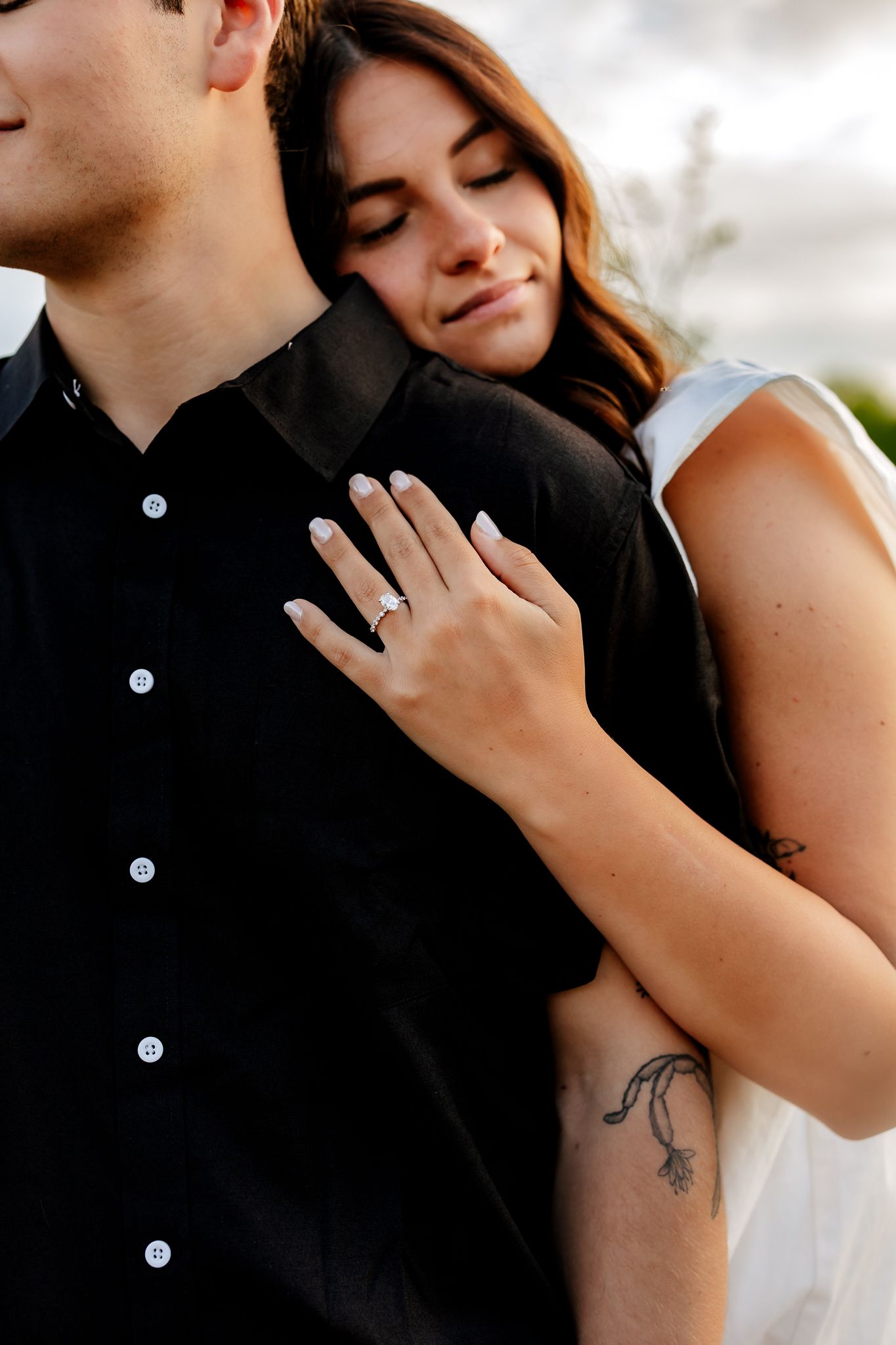 A woman with an engagement ring rests her hands on a mans chest in a tender close-up portrait outdoors.