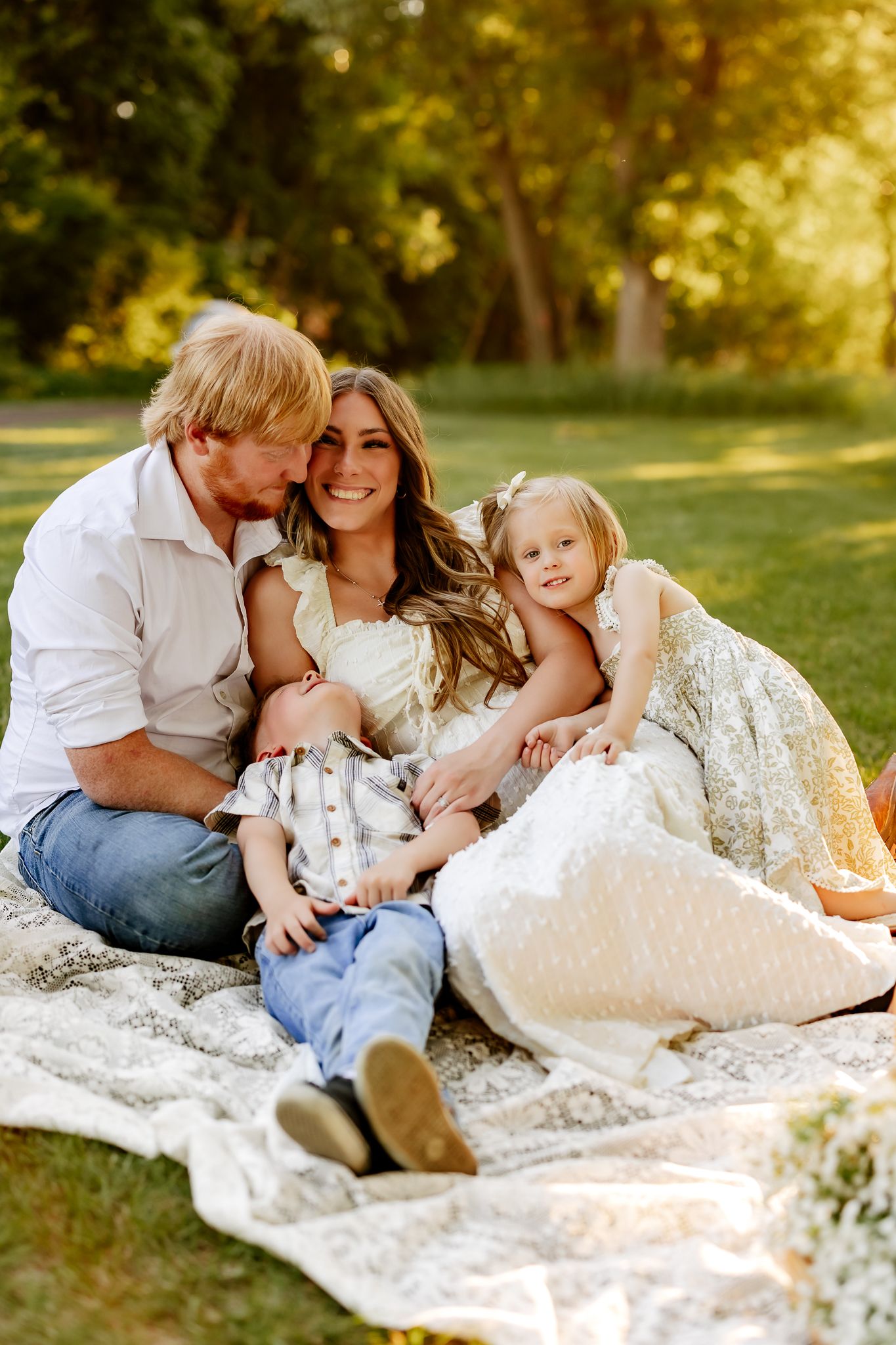 A smiling family of four sits together on a lace blanket in a sunlit park, sharing a warm embrace.