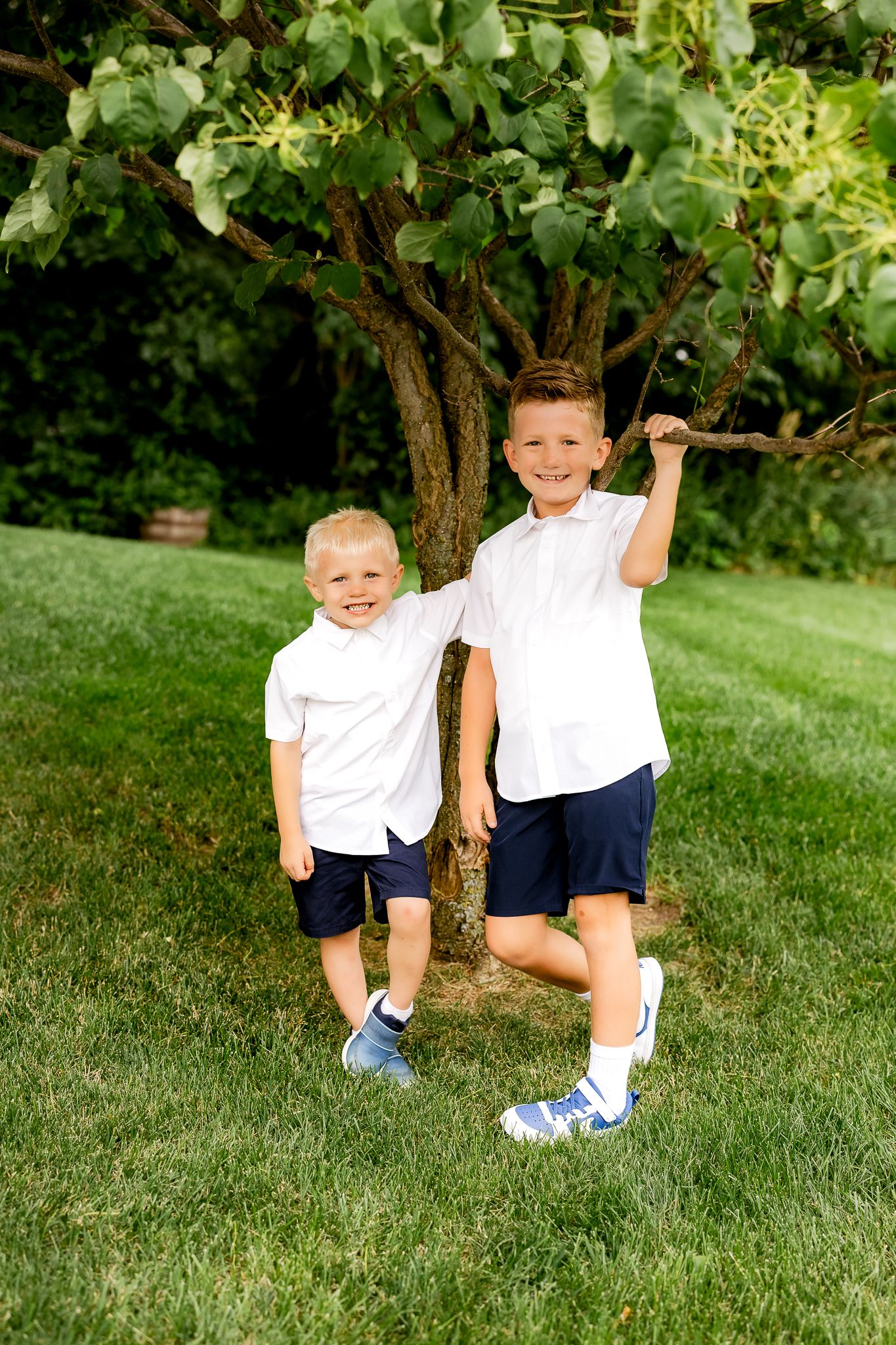 Two young boys in matching white shirts and navy shorts smile together outdoors near a tree on green grass.
