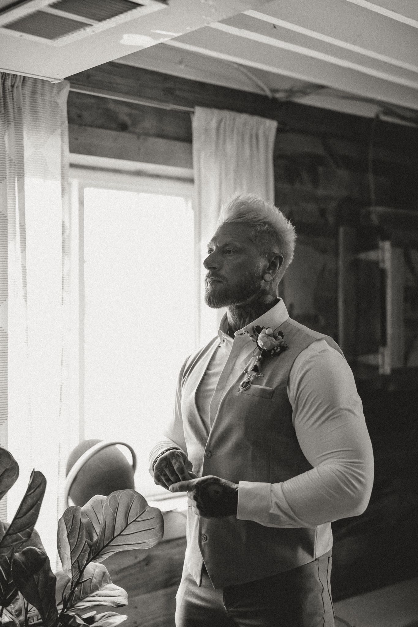 Black and white photo of a groom in a vest and boutonniere standing by a window on his wedding day.