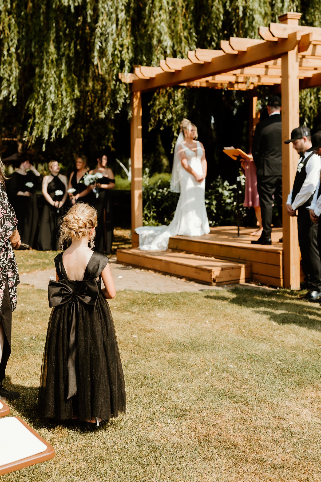 A flower girl in a black dress with bow watches an outdoor wedding ceremony under a wooden pergola surrounded by willow trees.