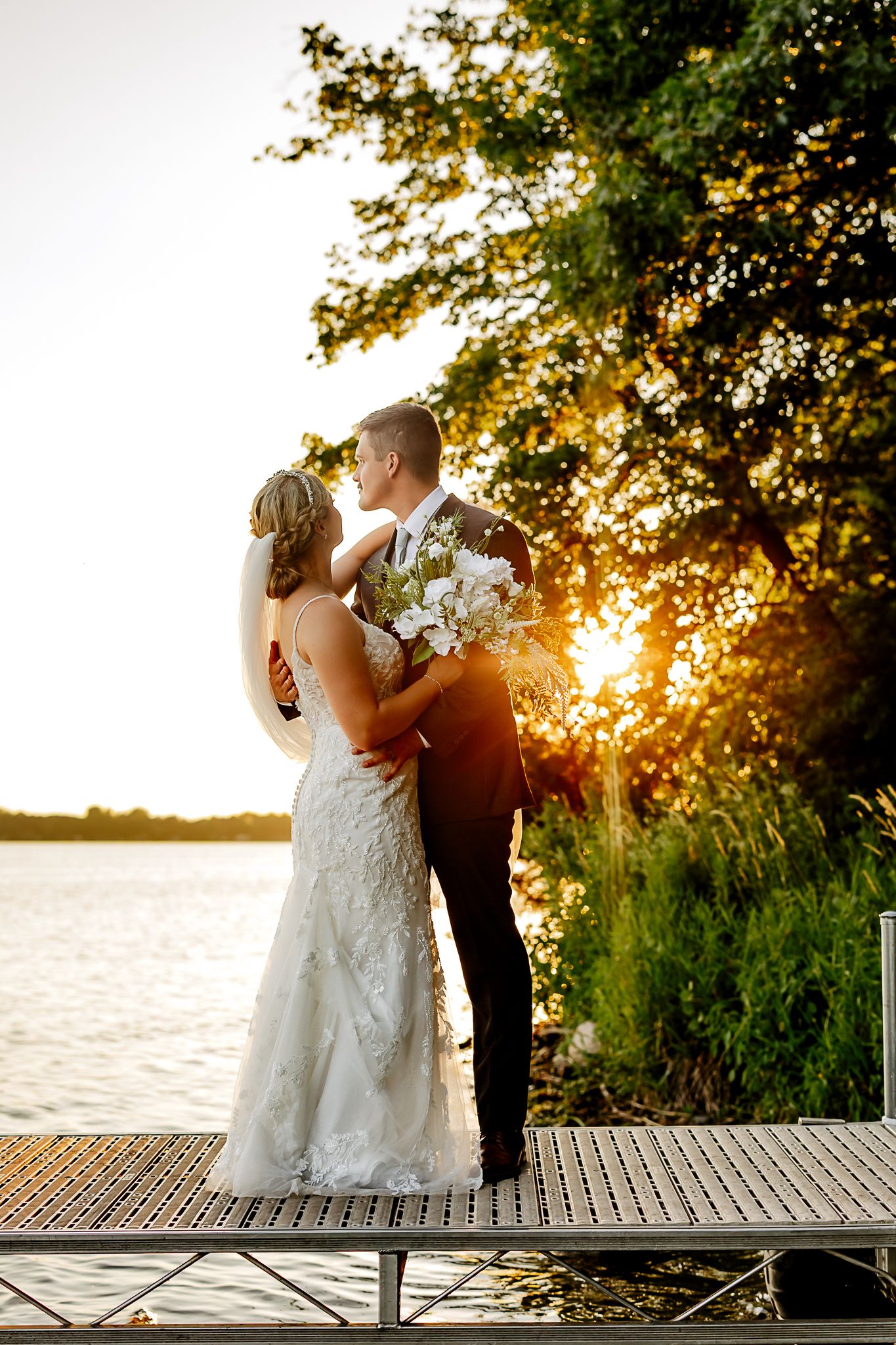 Bride and groom embrace on a lakeside dock at golden sunset, surrounded by lush trees, bride holding white bouquet.