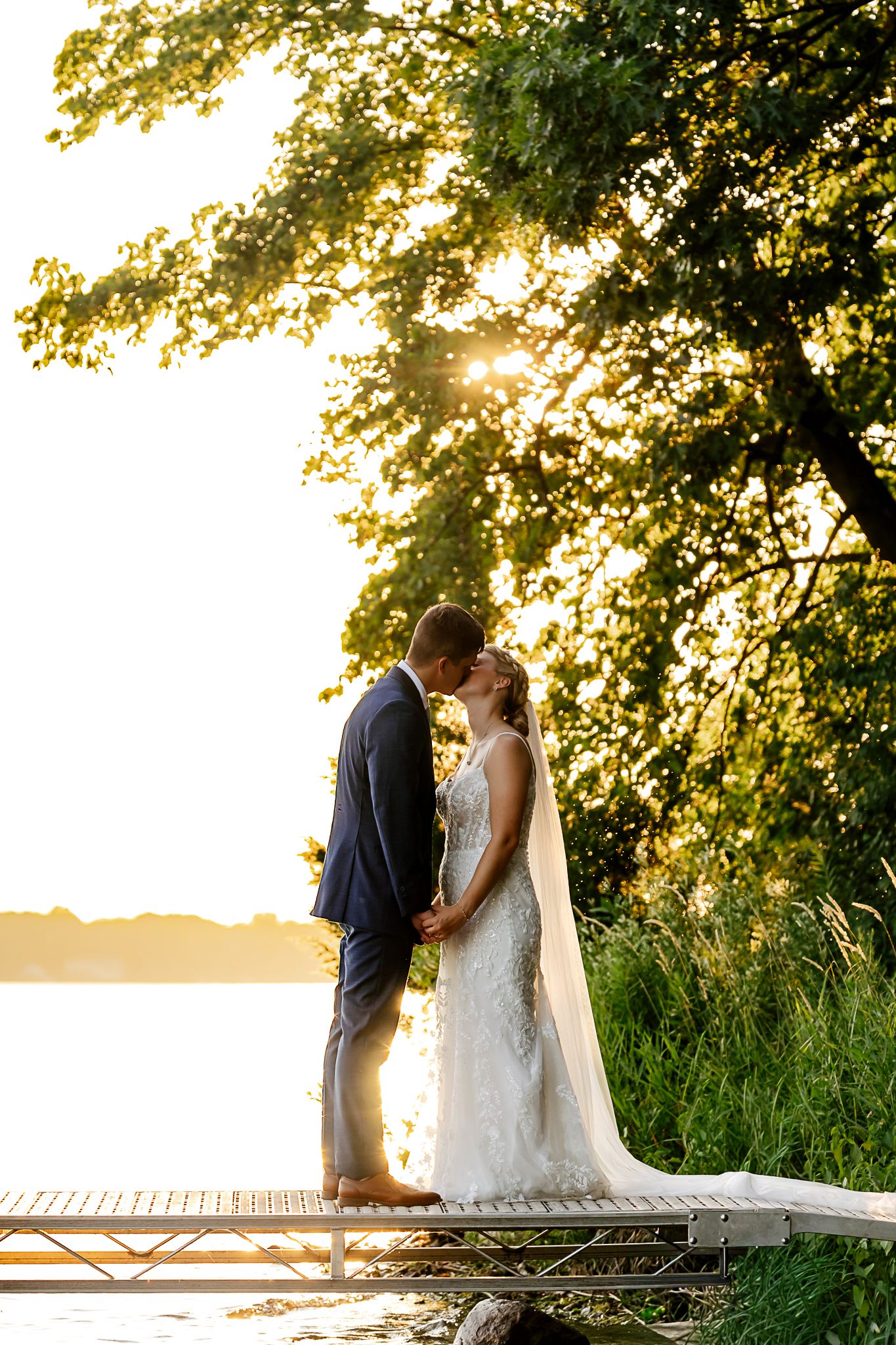 Bride and groom kissing on a dock at golden sunset by a lake, surrounded by lush green trees.