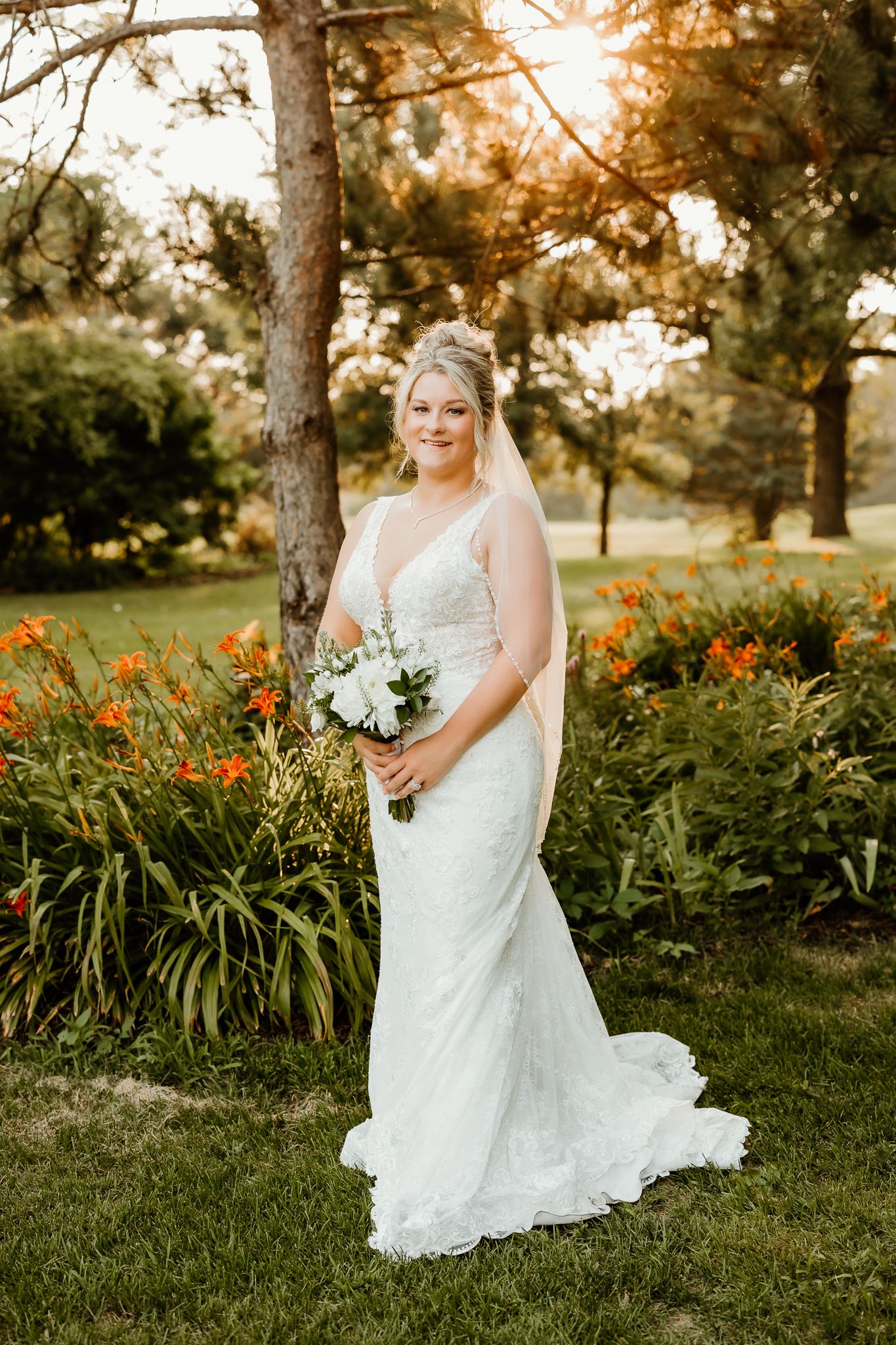 Bride in white lace gown holding bouquet stands in sunlit garden with orange flowers and trees.