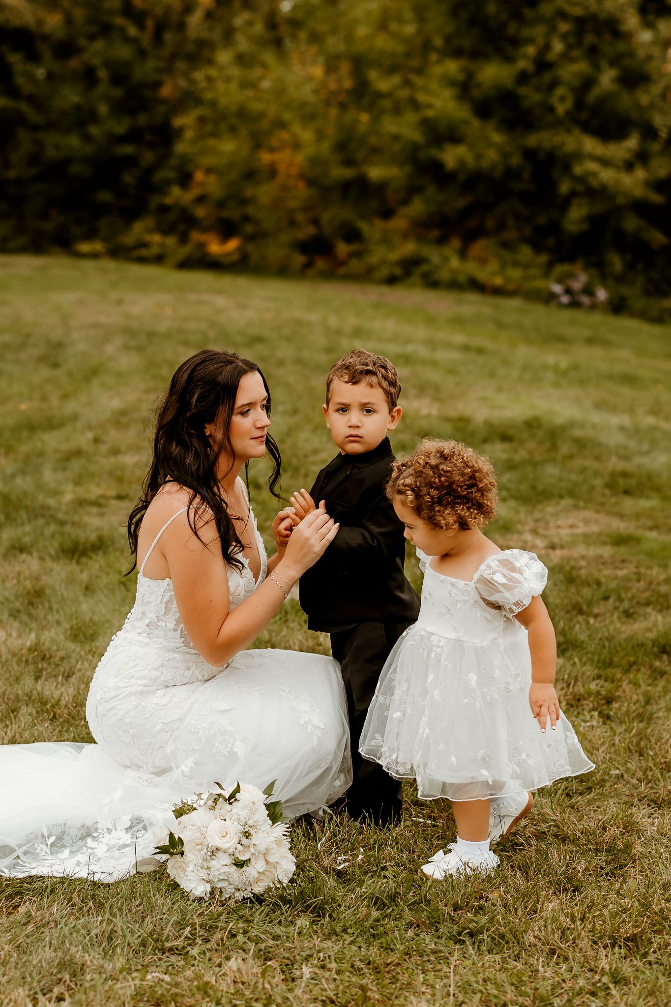 Bride in a white lace wedding gown kneels on grass beside a boy in a black suit and a flower girl in a white tulle dress.